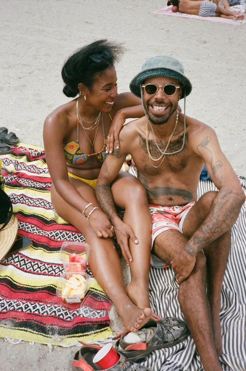 A smiling woman and a man sitting closely on a beach blanket, enjoying a sunny day. The woman is wearing a colorful bikini, and the man is shirtless in shorts, with sunglasses and a bucket hat. They are surrounded by beach accessories and food.