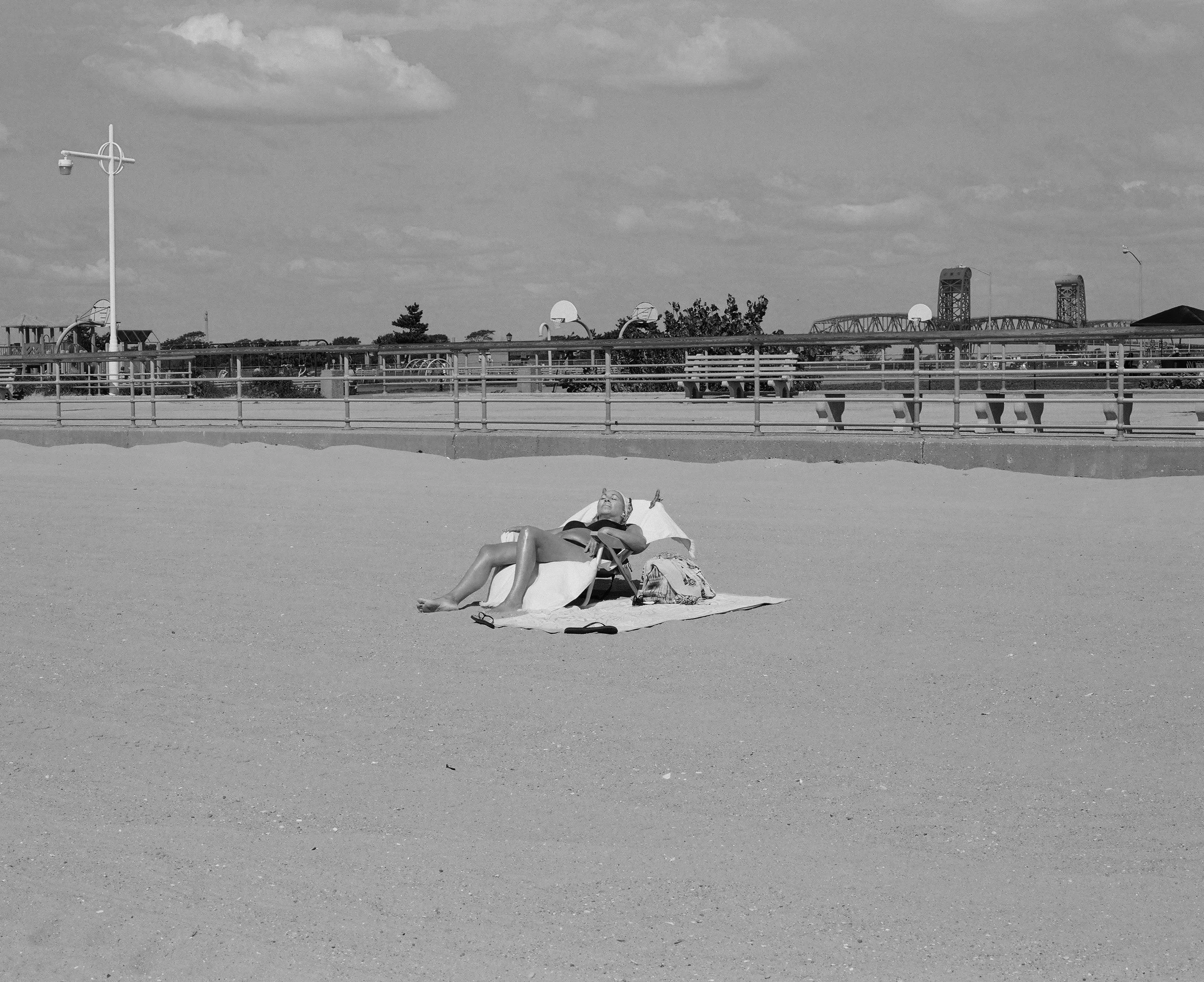A black and white photo of a person relaxing on a lounge chair in a park or playground, with a fence and playground equipment in the background, under a partly cloudy sky.