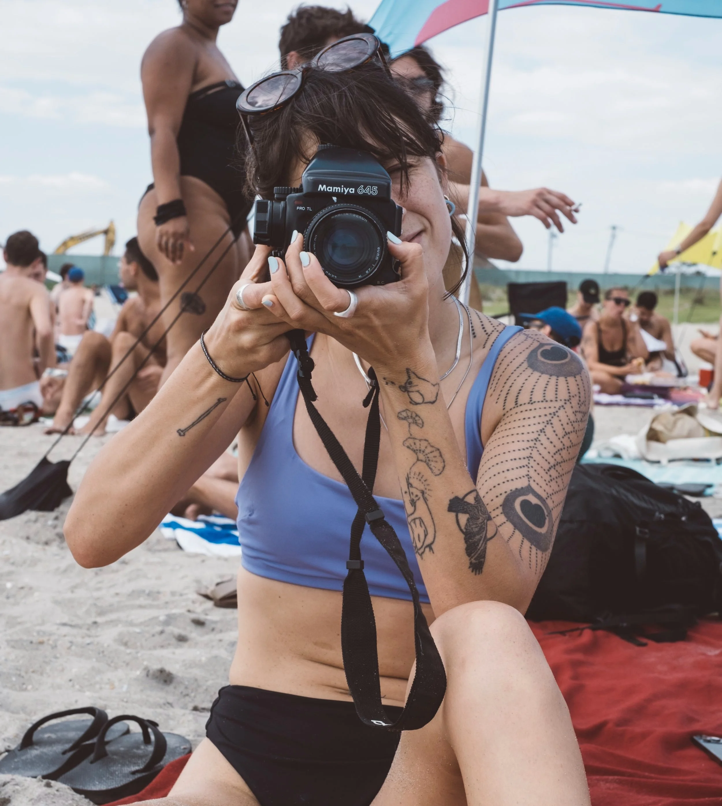 A woman at the beach taking a photo with a vintage Mamiya 645 camera. She is seated on the sand, wearing a blue tank top, black shorts, and sunglasses on her head. Several people are in the background, some sitting and others standing, with umbrellas and beach gear around.
