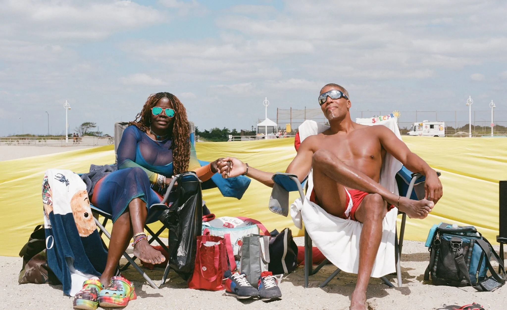 Two people sitting outdoors on beach chairs, holding hands. The woman has long, curly hair and is wearing sunglasses, a blue sheer top, and colorful shoes. The man is shirtless, wearing red swim trunks, sunglasses, and has a beard. There is beach gear and bags around them, and a yellow barrier in the background with a partly cloudy sky.