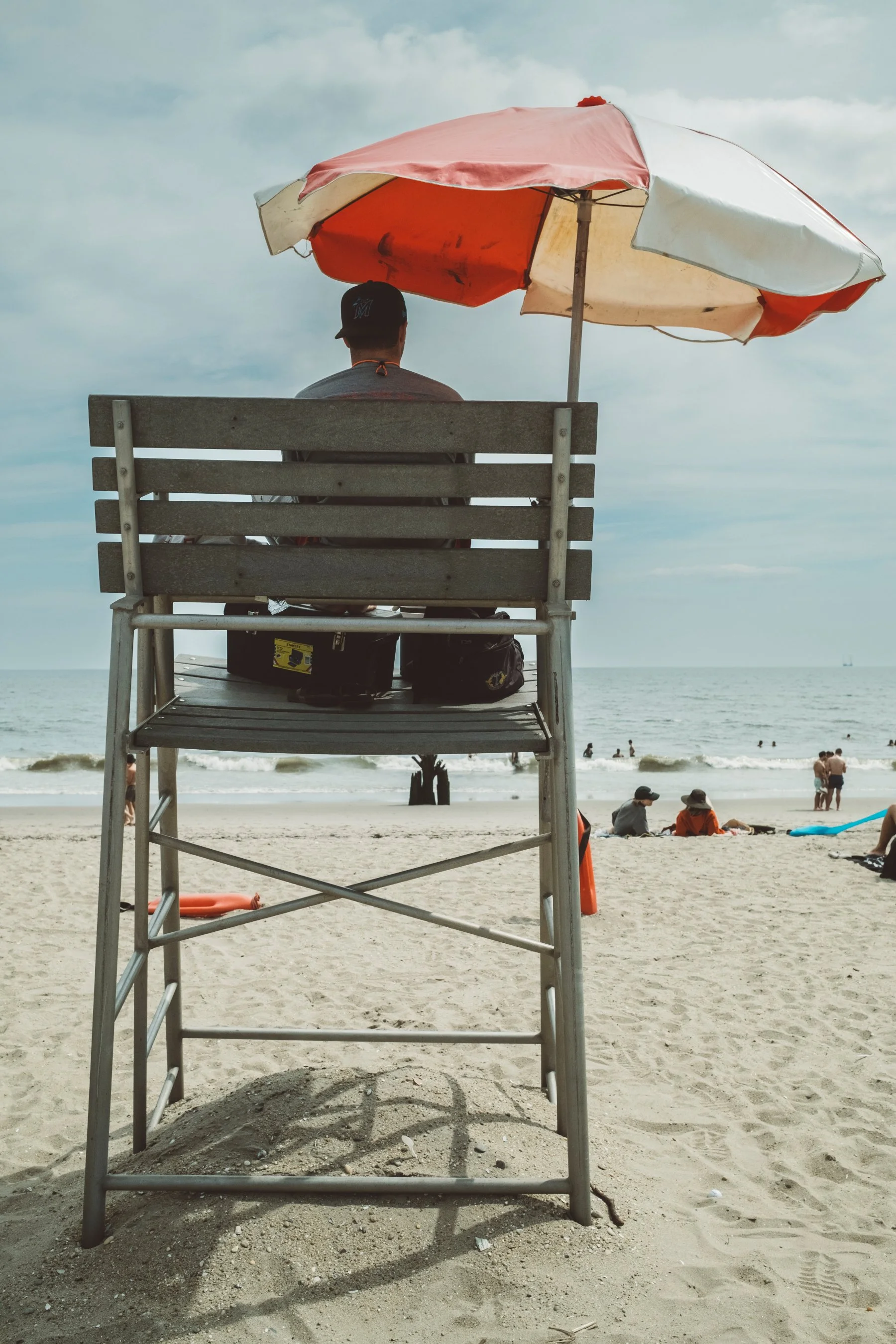 A person sitting on a lifeguard chair beneath a large red and white beach umbrella at the beach, with the ocean and people in the background.