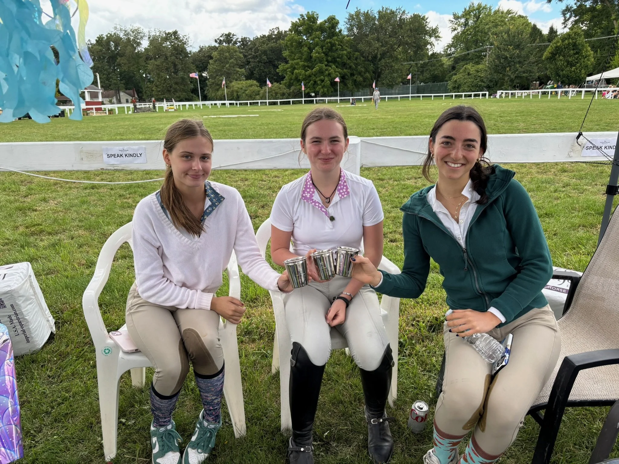 Three young women sitting on white chairs on grass at an outdoor event, holding metallic cups and smiling. In the background, there is a racecourse with flags, a white fence, and a person riding a horse.