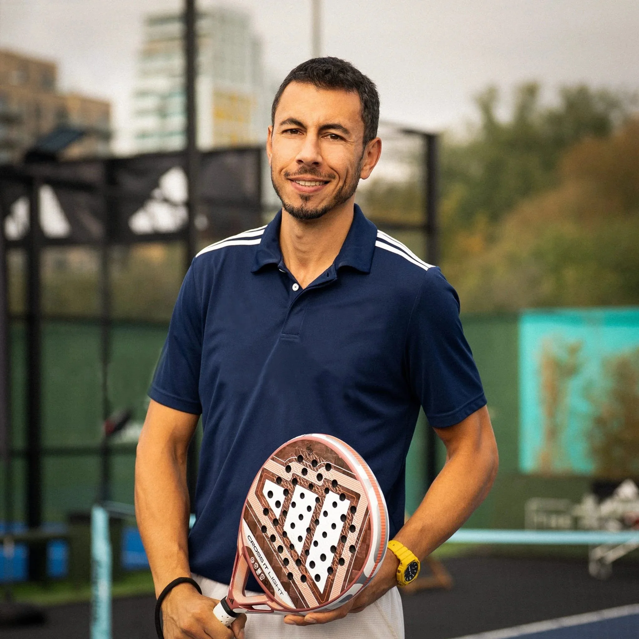 A player in a black shirt and white shorts serves a ball on a blue paddle court by the beach, with straw umbrellas and blue ocean in the background.