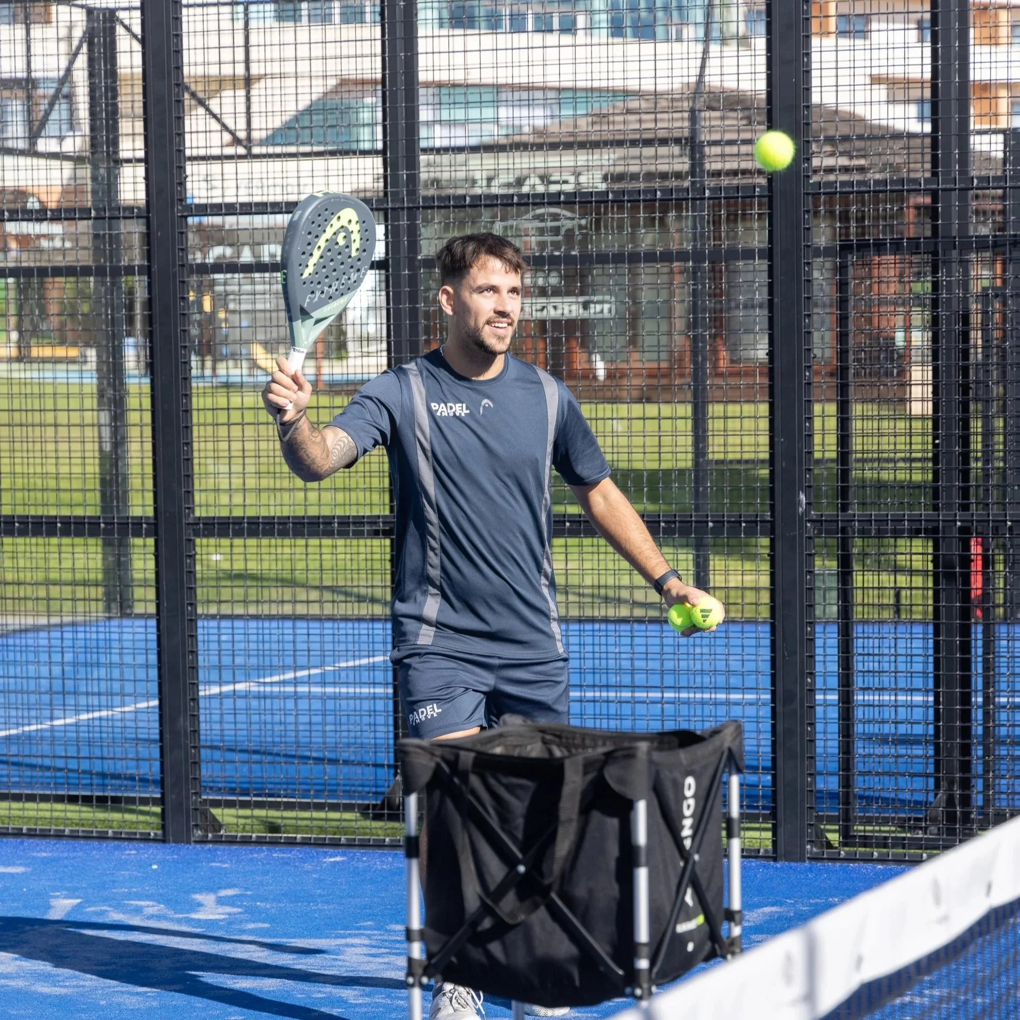A padel coach feeding balls to a participant during the padel training camp.  The two men are on a blue court, surrounded by a wire fence, with padel rackets and padel balls visible.