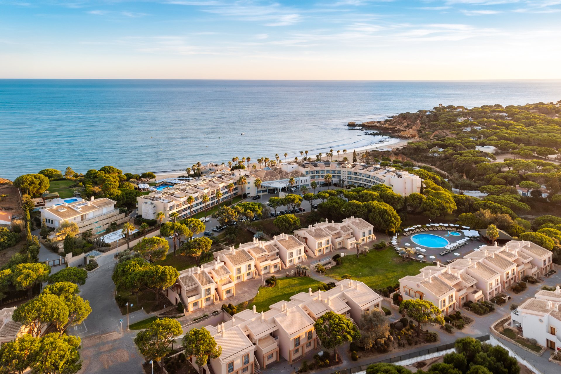 Aerial view of a coastal resort with white buildings, swimming pools, and lush green trees, overlooking the ocean on a clear day.