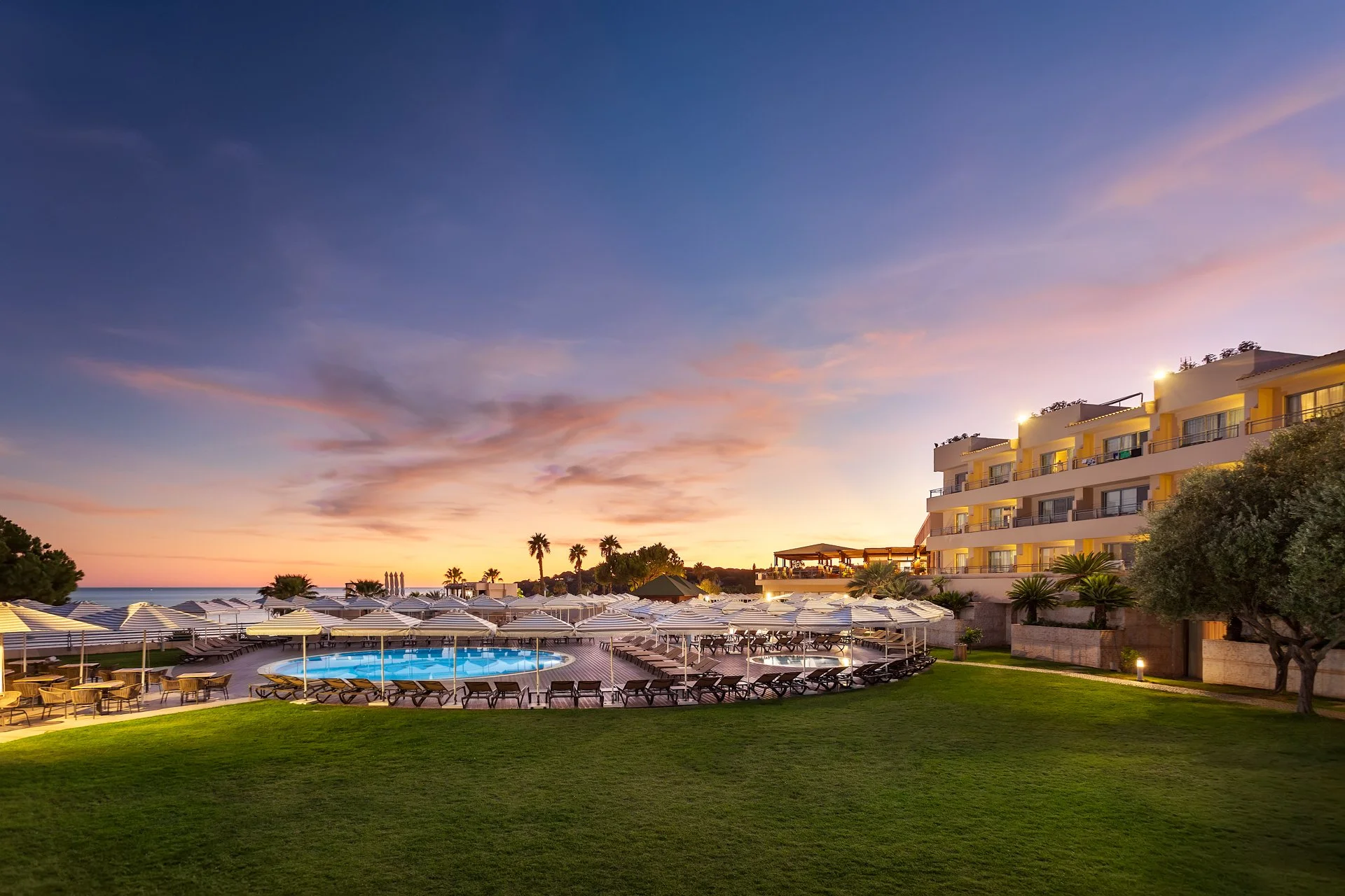 Sunset view of a seaside resort with swimming pool, numerous white umbrellas and loungers, multi-story yellow and white hotel building, palm trees, and a cloudy sky.