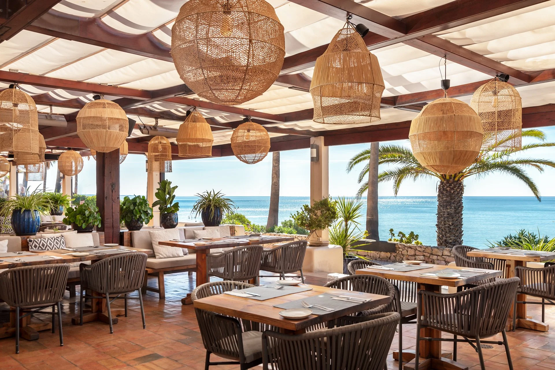 An outdoor seaside restaurant with wooden furniture, potted plants, and woven pendant lights, overlooking a beach and ocean.