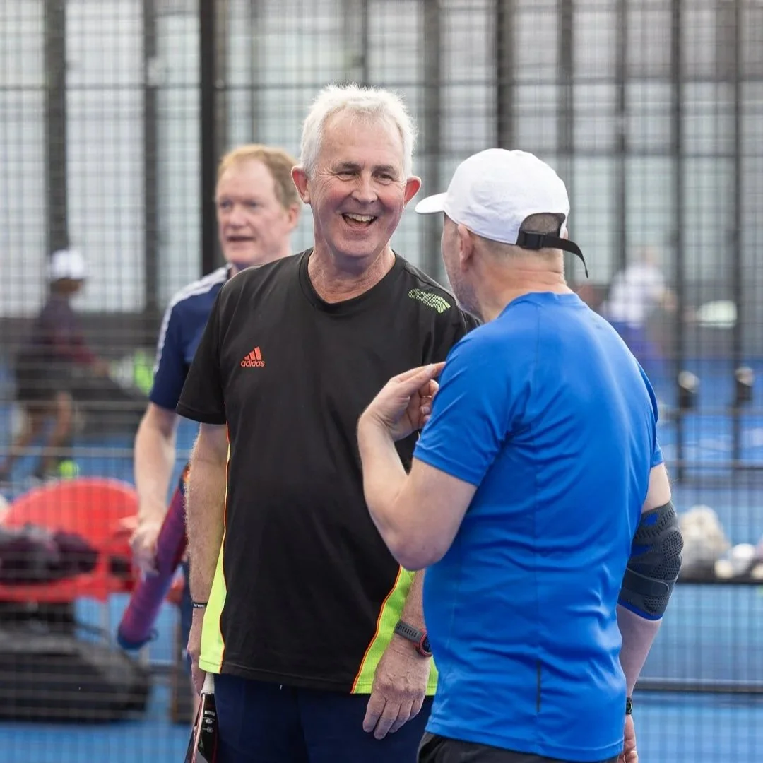 A padel coach feeding balls to a participant during the padel training camp. The two men are on a blue court, surrounded by a wire fence, with padel rackets and padel balls visible.