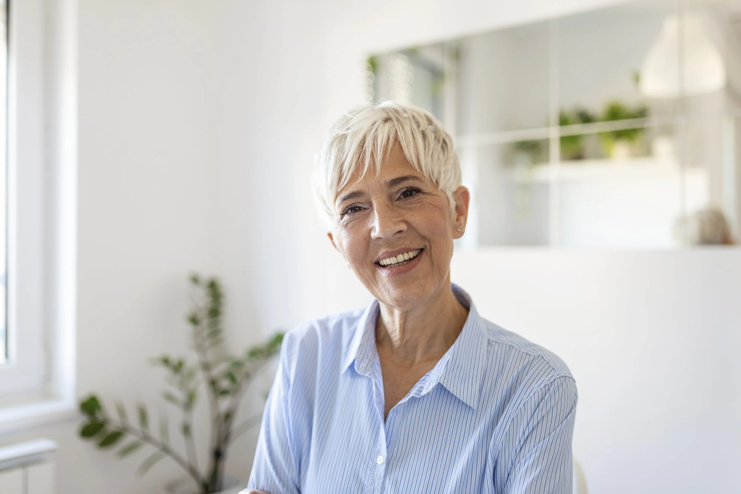 A smiling older woman with short blonde hair wearing a light blue striped shirt, sitting in a bright room with white walls and a window, and some green plants in the background.