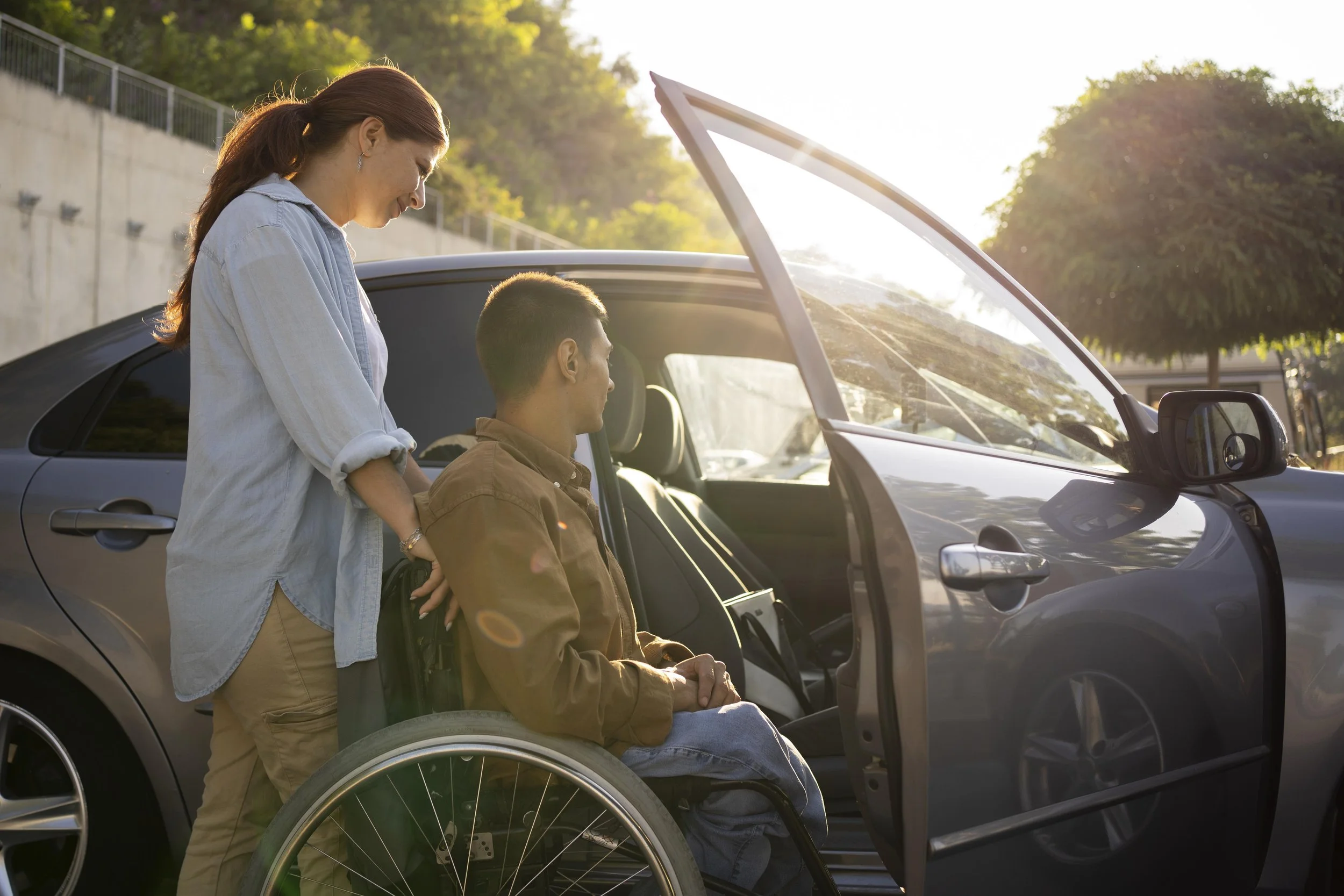 A woman standing behind a man in a wheelchair outside a car, on a sunny day.