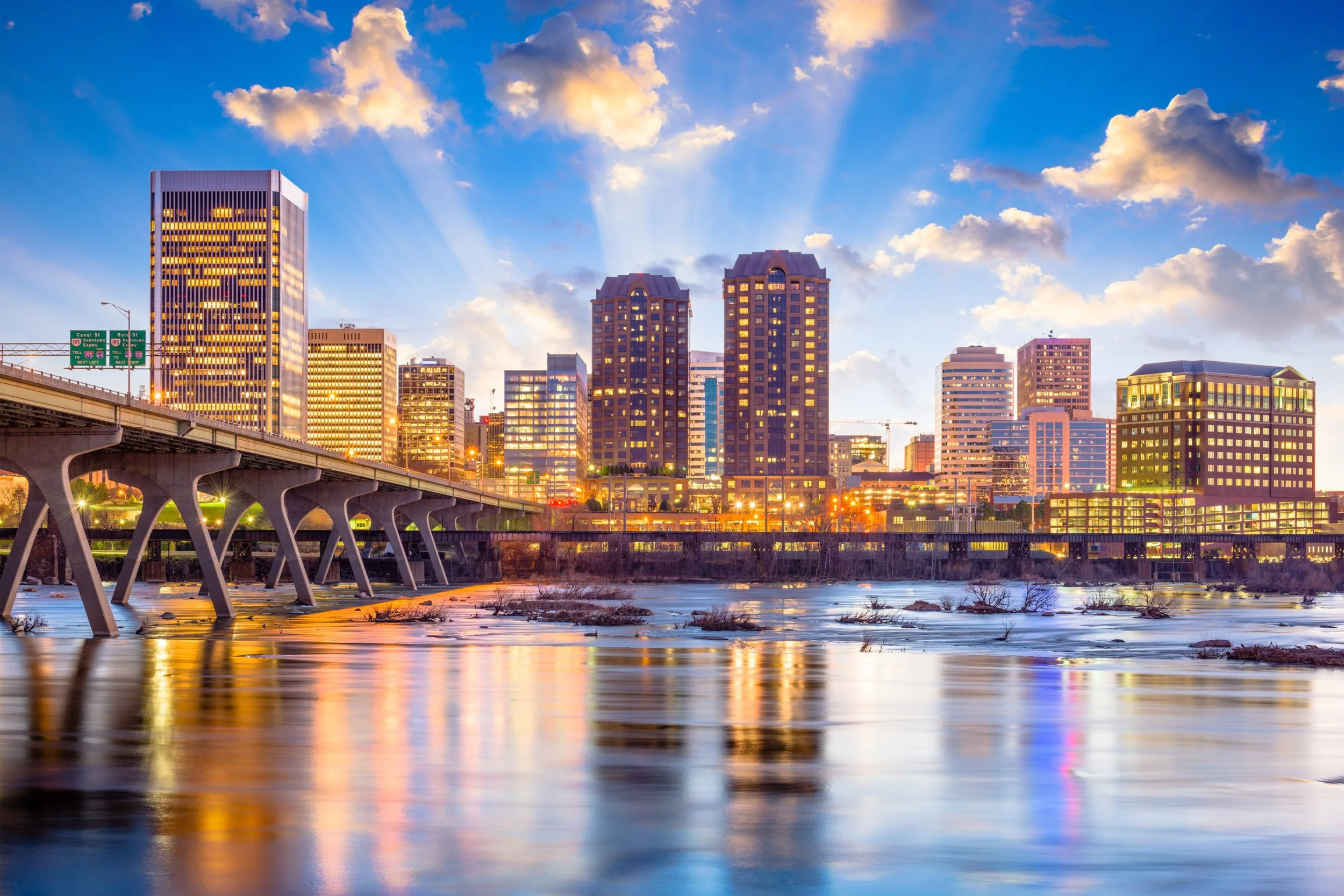City skyline at sunset with tall buildings, a bridge over a river, and clouds in the sky.