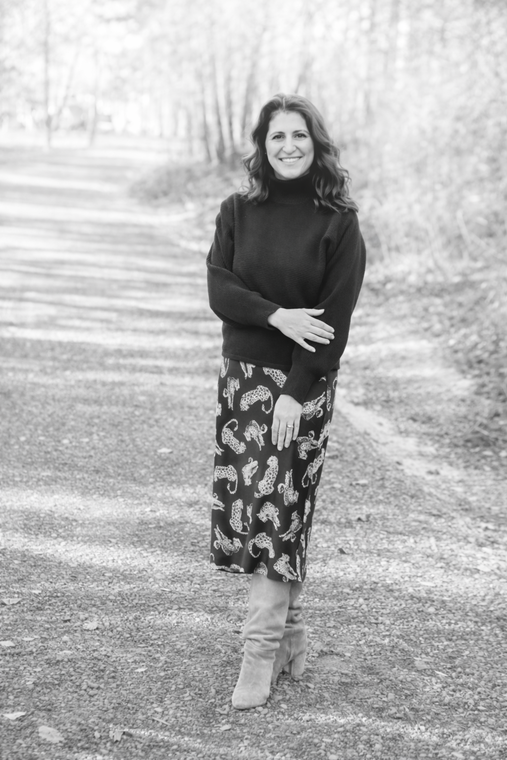 A woman smiling outdoors in black and white, wearing a dark turtleneck sweater, a leopard print skirt, and high-heeled boots, standing on a dirt path surrounded by trees.