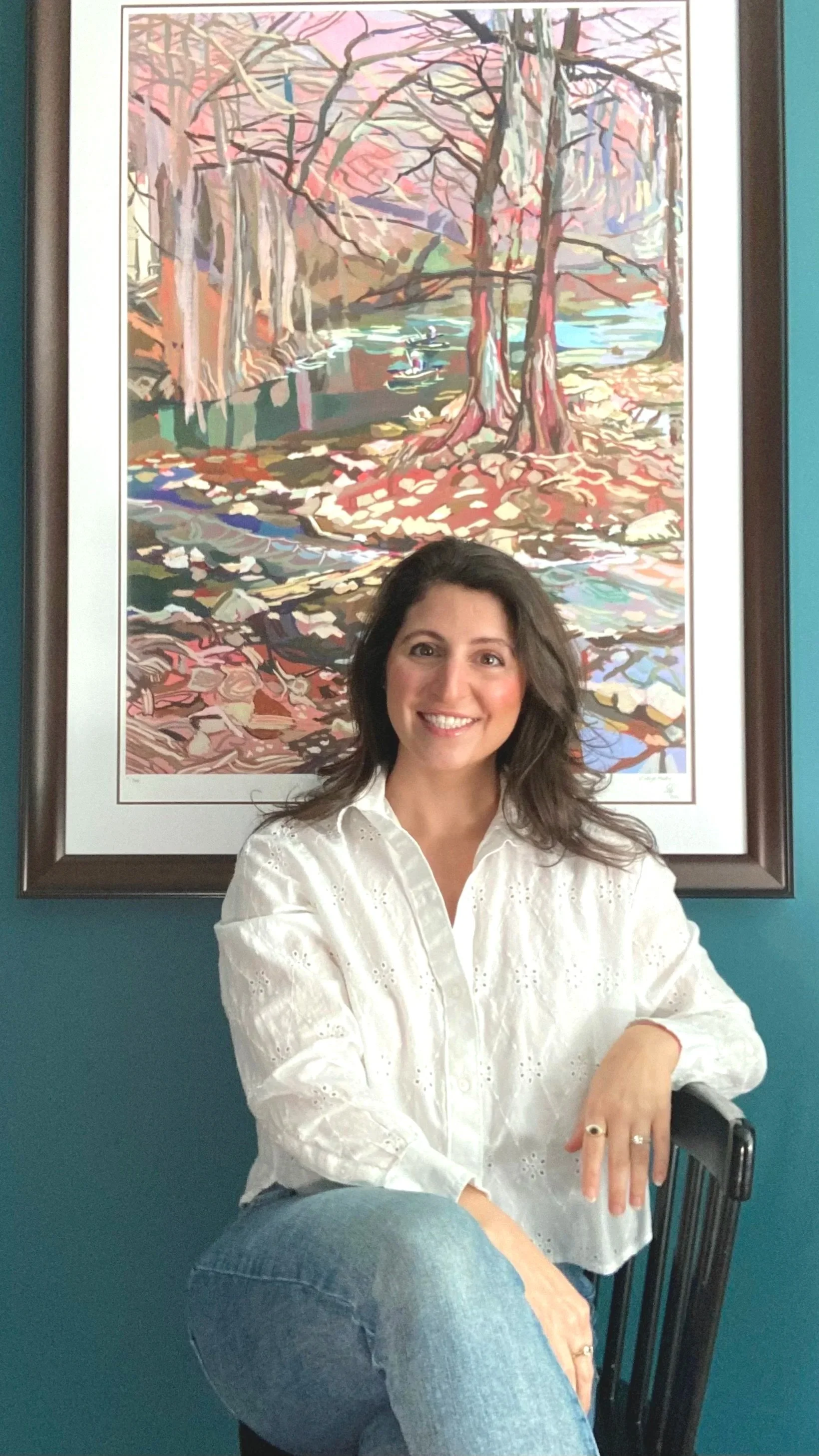 A smiling woman with dark brown hair, wearing a white eyelet blouse and blue jeans, sitting on a chair in front of a teal wall. Behind her is a painting of a colorful scene of trees and a creek.