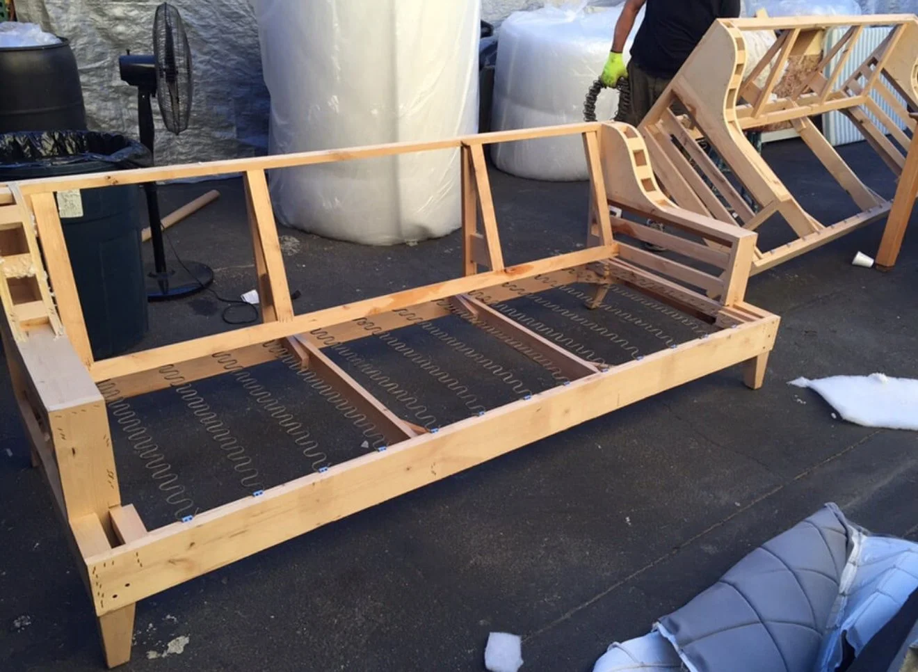 Wooden couch frame under construction outdoors, with metal springs visible and cushions nearby. Person in gloves working in the background.