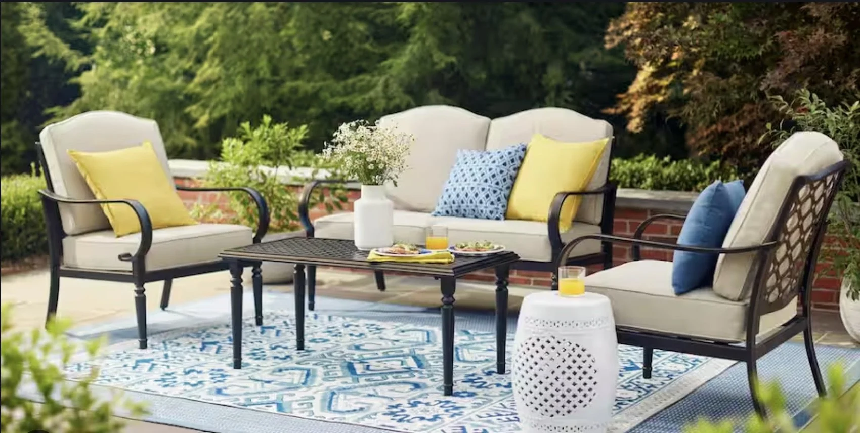 Outdoor patio furniture set with a sofa, two chairs, and a table on a blue patterned rug. The sofa has yellow and blue cushions. There's a white vase with flowers on the table and two plates with food accompanied by glasses of juice.
