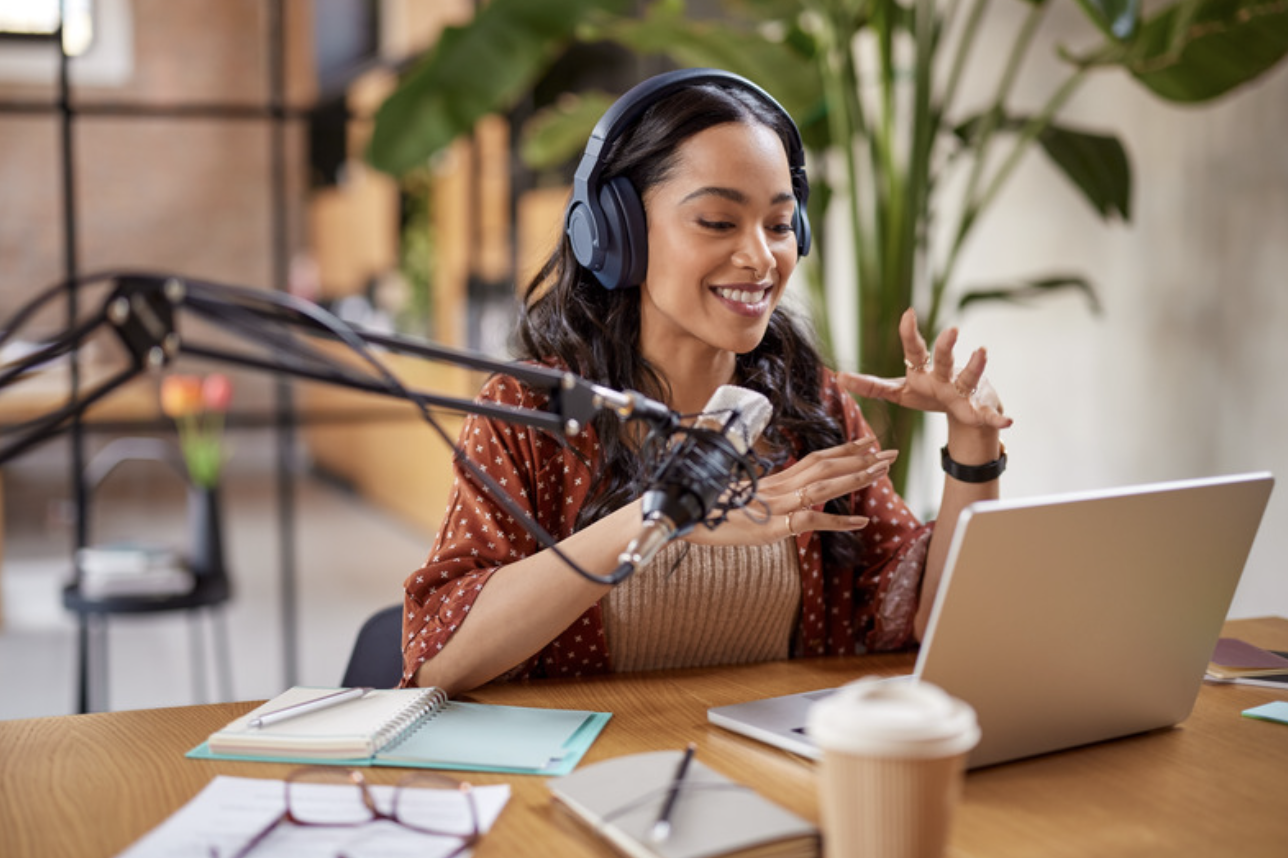 Woman recording podcast with laptop and microphone, wearing headphones, in a modern office setting.