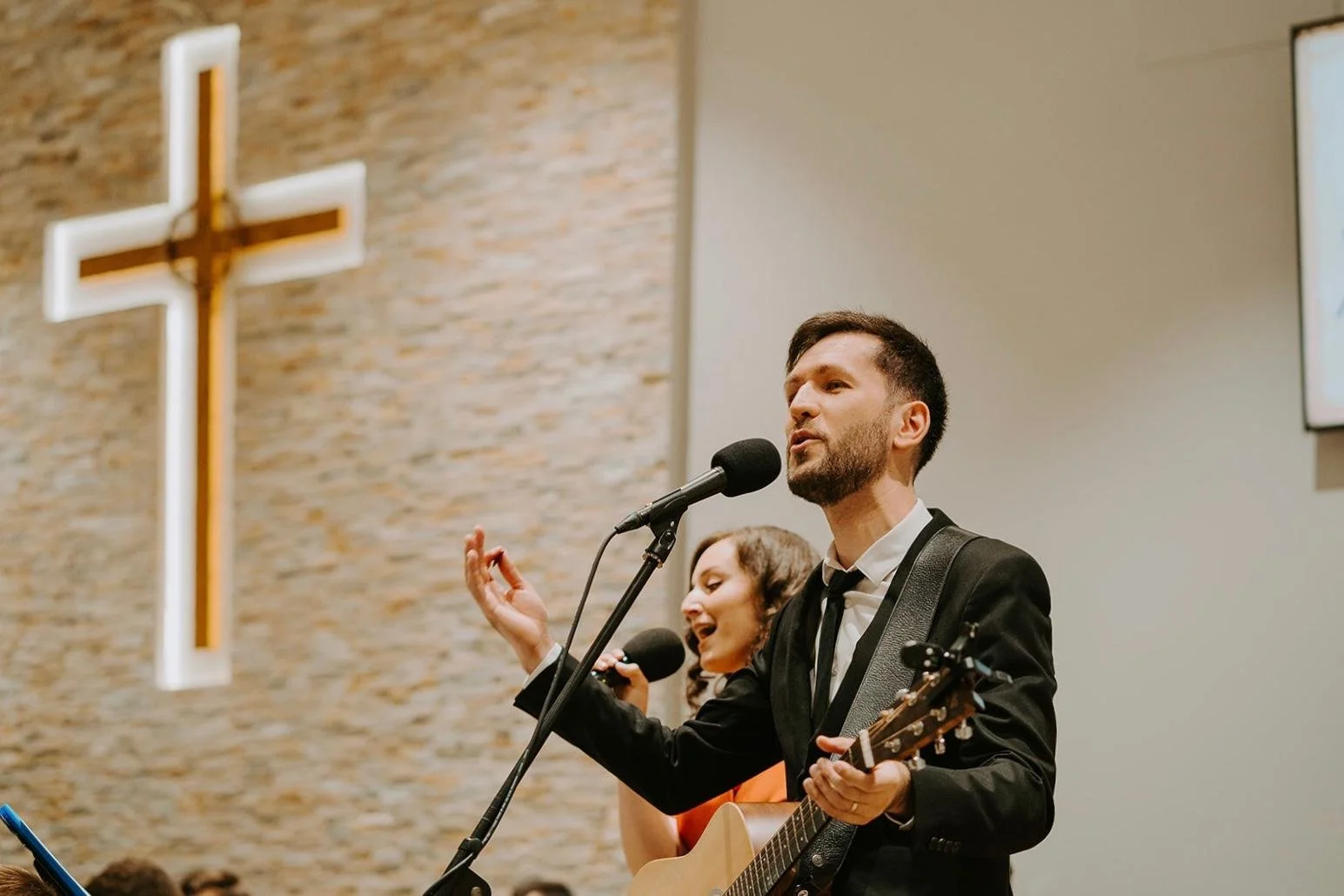 A man in a black suit playing guitar and singing into a microphone during a church service or concert, with a woman singing and holding a microphone in the background, inside a church with a large illuminated cross on the brick wall.