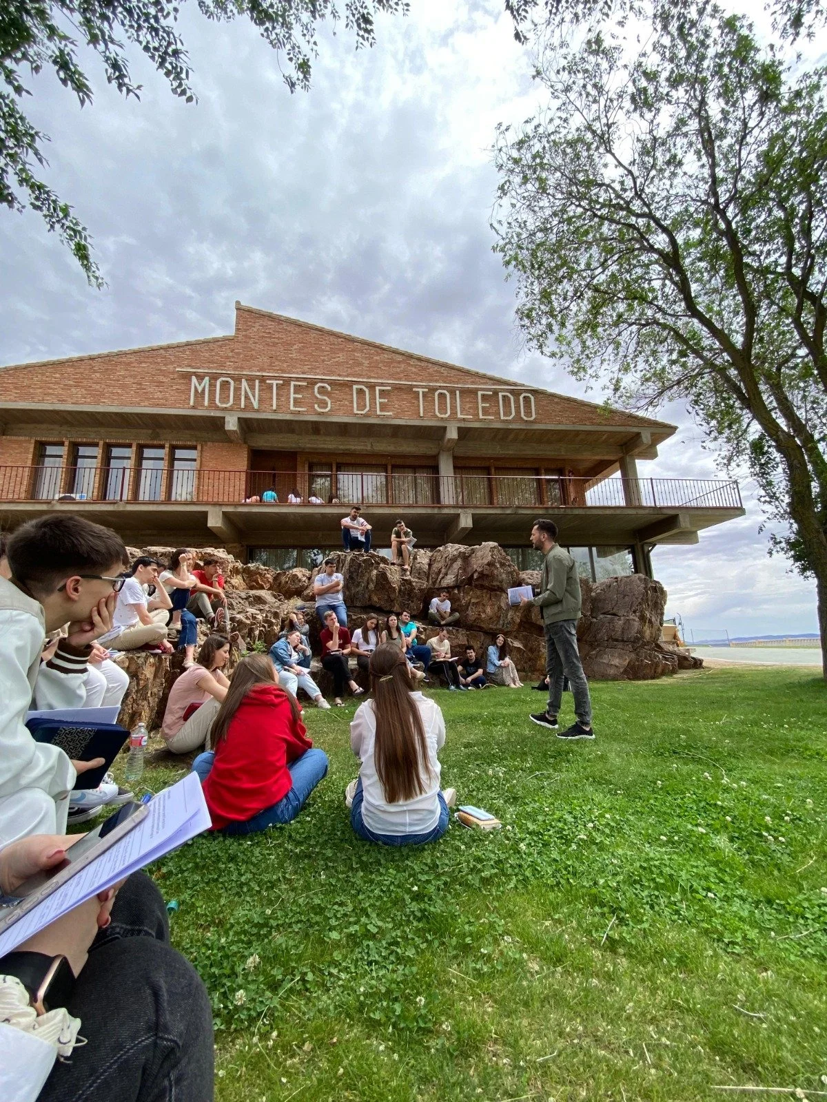 Group of students attending an outdoor class or lecture at Montes de Toledo, sitting on rocks and grass near a modern building with the name "Montes de Toledo." A man is standing and speaking to the group, holding a notebook, on cloudy day with trees