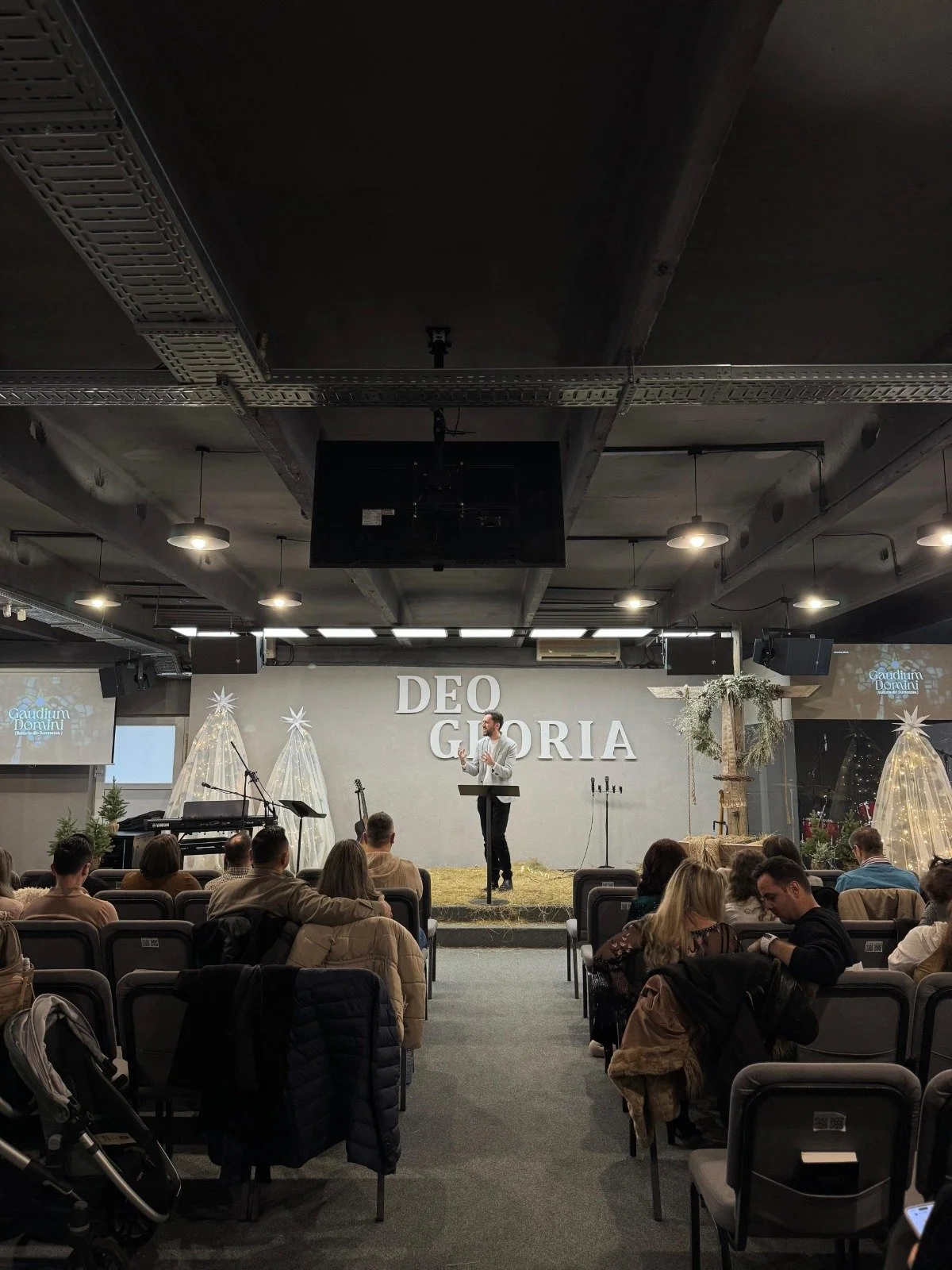 A man in a white jacket giving a speech on stage during a church service or event at Deus Gloria. The stage is decorated with Christmas trees and festive decorations. There are people seated and listening, and a few chairs and musical instruments on 