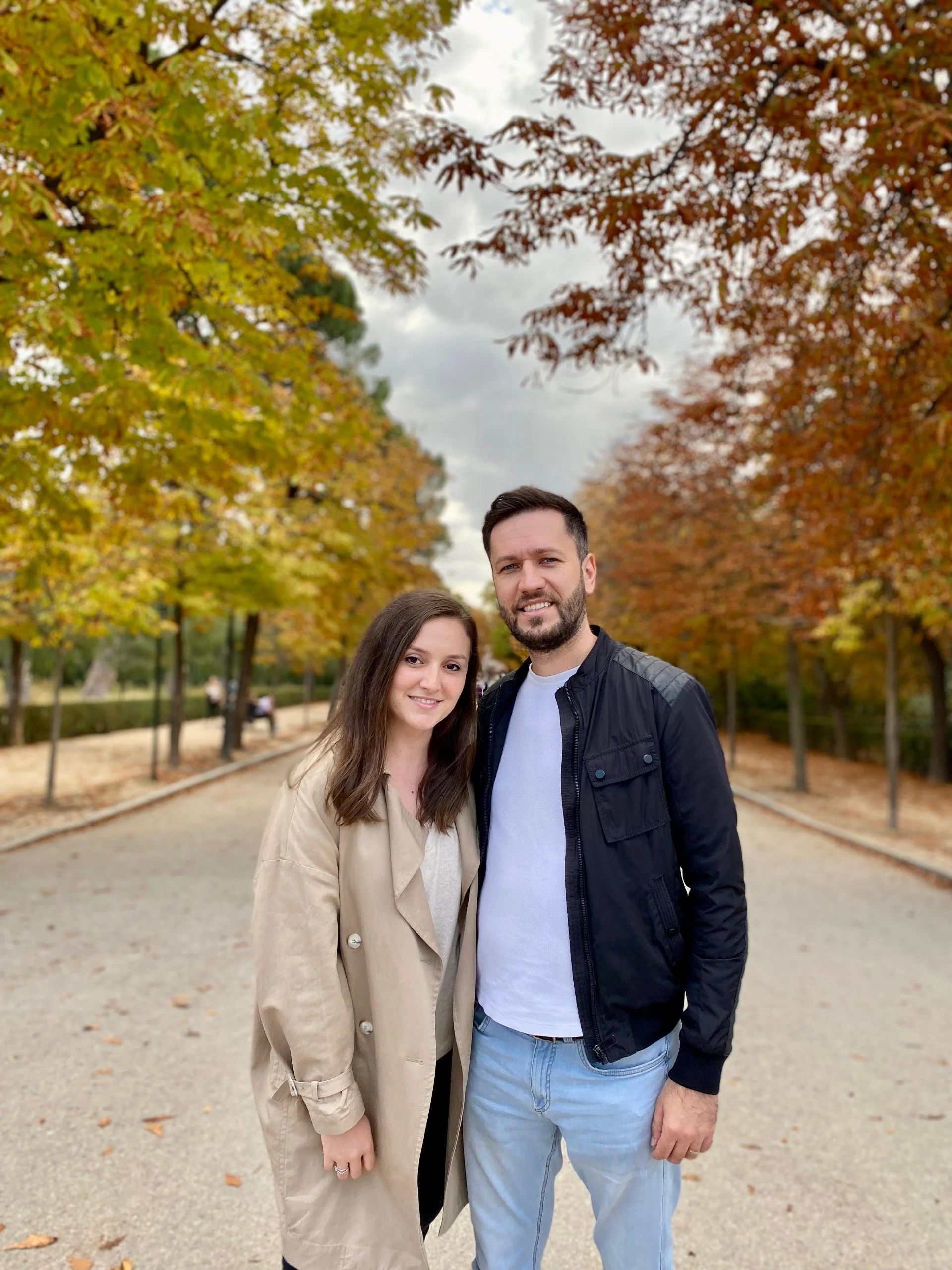 A man and woman standing outdoors on a tree-lined pathway during fall, with colorful autumn leaves on the trees and some fallen on the ground.