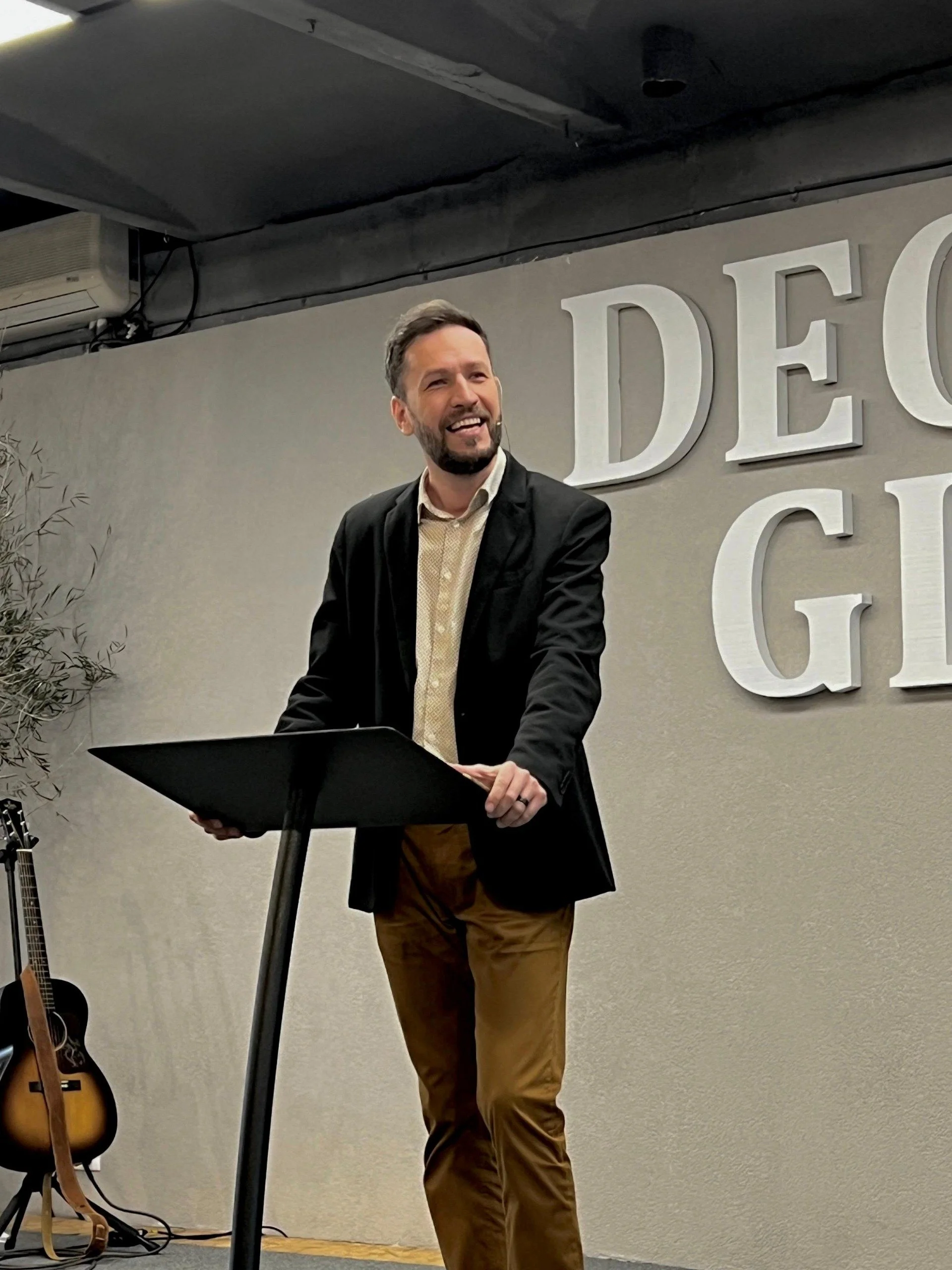 A man with a beard and mustache, wearing a black blazer and tan pants, is standing behind a music stand, smiling and speaking. Behind him is a wall with large white letters spelling 'DECO.' to the right, and a guitar leaning against the wall on the l