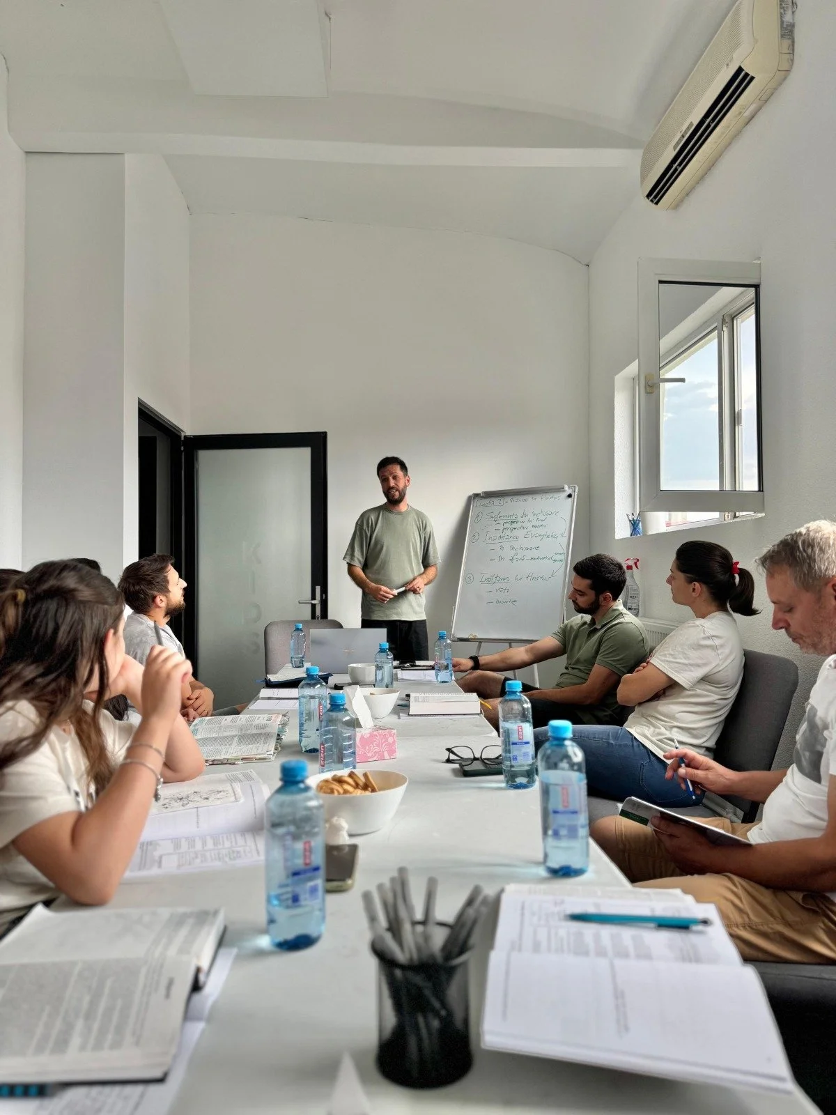 A man giving a presentation in a conference room with eight people seated around a table, several bottles of water, books, and personal items on the table.