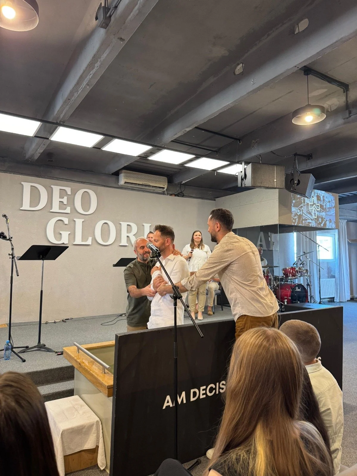 People on stage at a church or event space, with the words "DEO GLORIA" on the wall, participating in a ceremony or presentation, with musical instruments in the background and an audience watching.