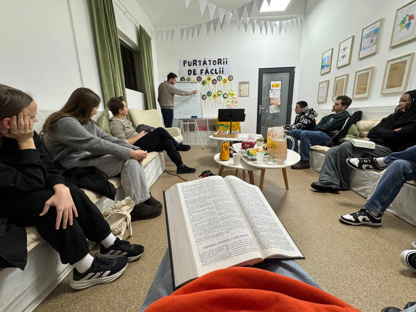A group of people attending a casual indoor meeting or class, sitting on sofas and chairs in a room with art on the walls. A man is standing near a whiteboard, writing or presenting. There are tables with snacks, drinks, and a large open book in the 