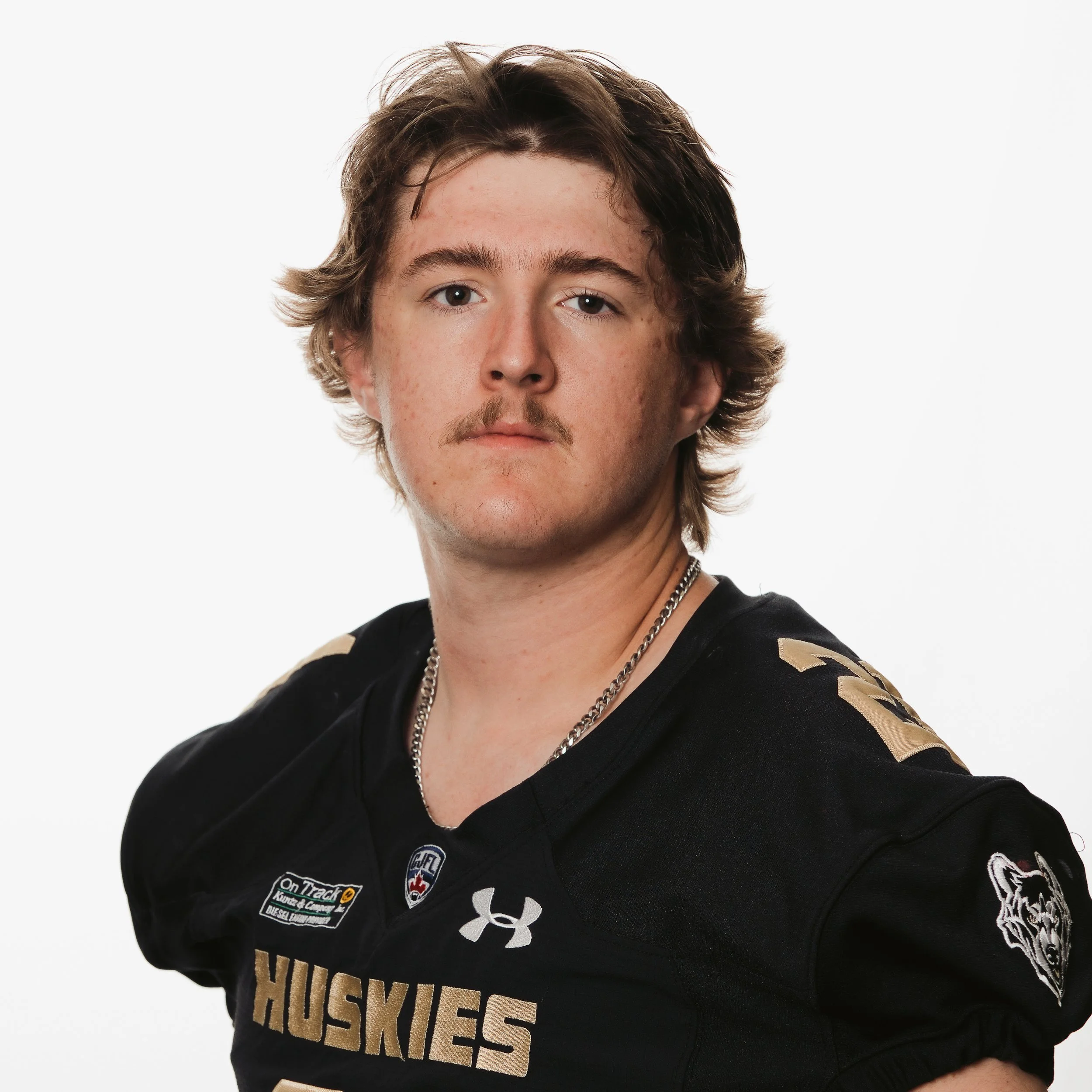 Young male football player in black Husky jersey with gold numbers and text, standing with arms behind back, looking at camera.
