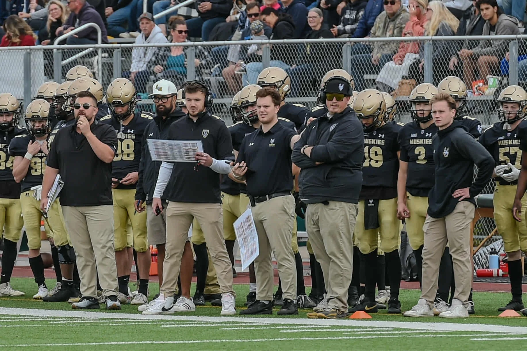 Group of football coaches and players standing outdoors, with coaches wearing jackets and sunglasses, and players in football helmets and uniforms.