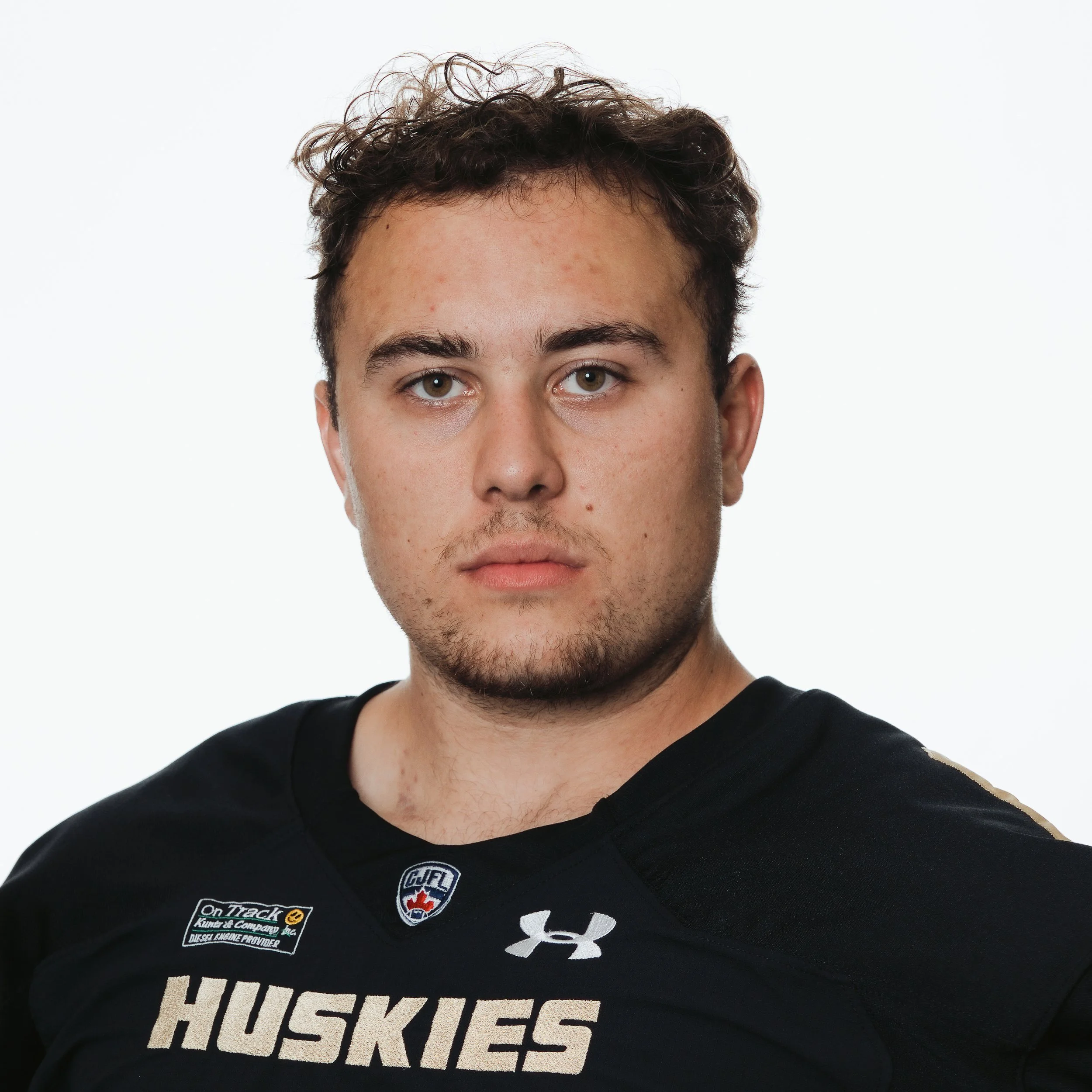 Close-up of a young man with curly brown hair wearing a black sports jersey with the word 'Huskies' and various patches, including an Under Armour logo and a Canadian flag emblem.