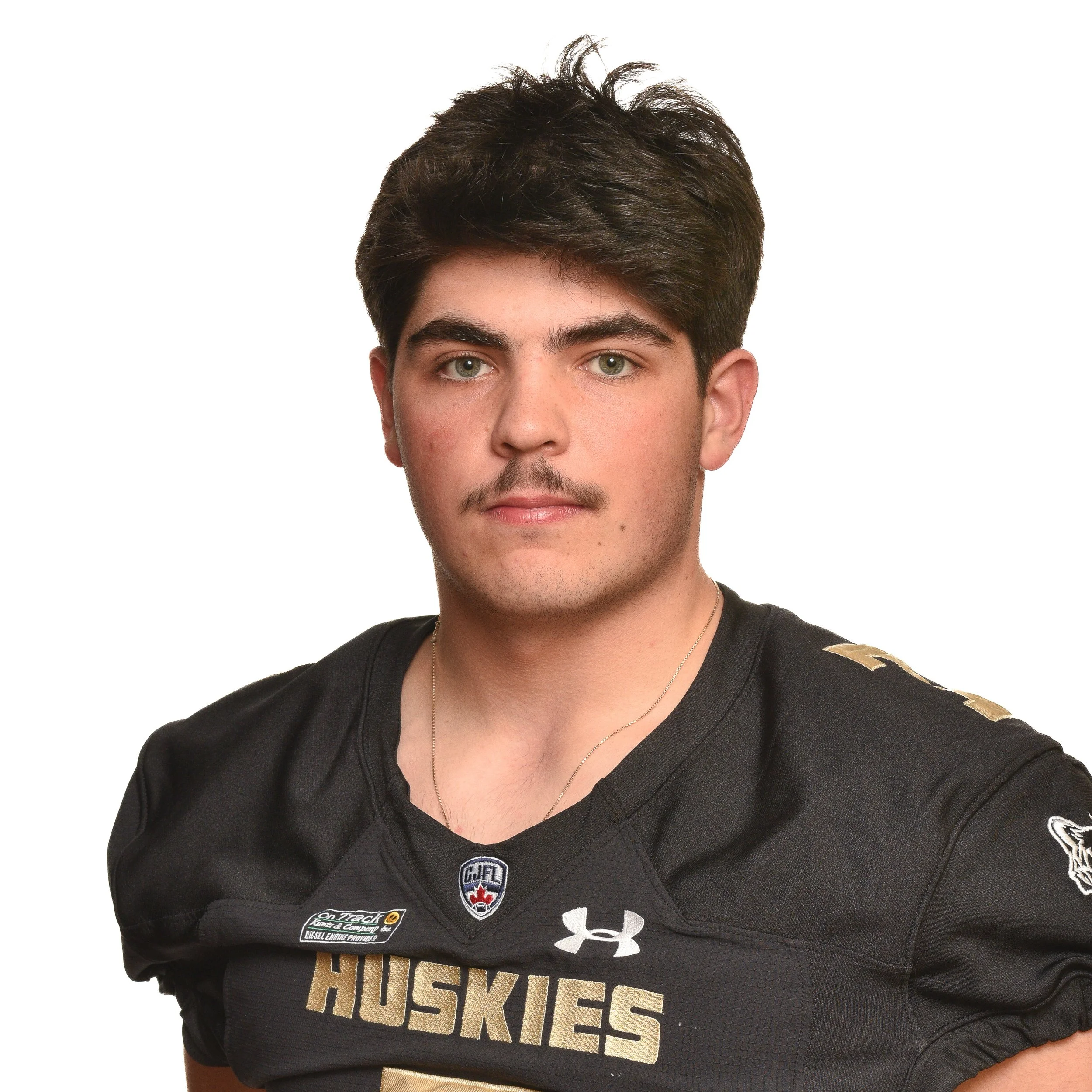 A young man with dark hair, wearing a black football jersey with 'Huskies' written on it, standing against a black background.