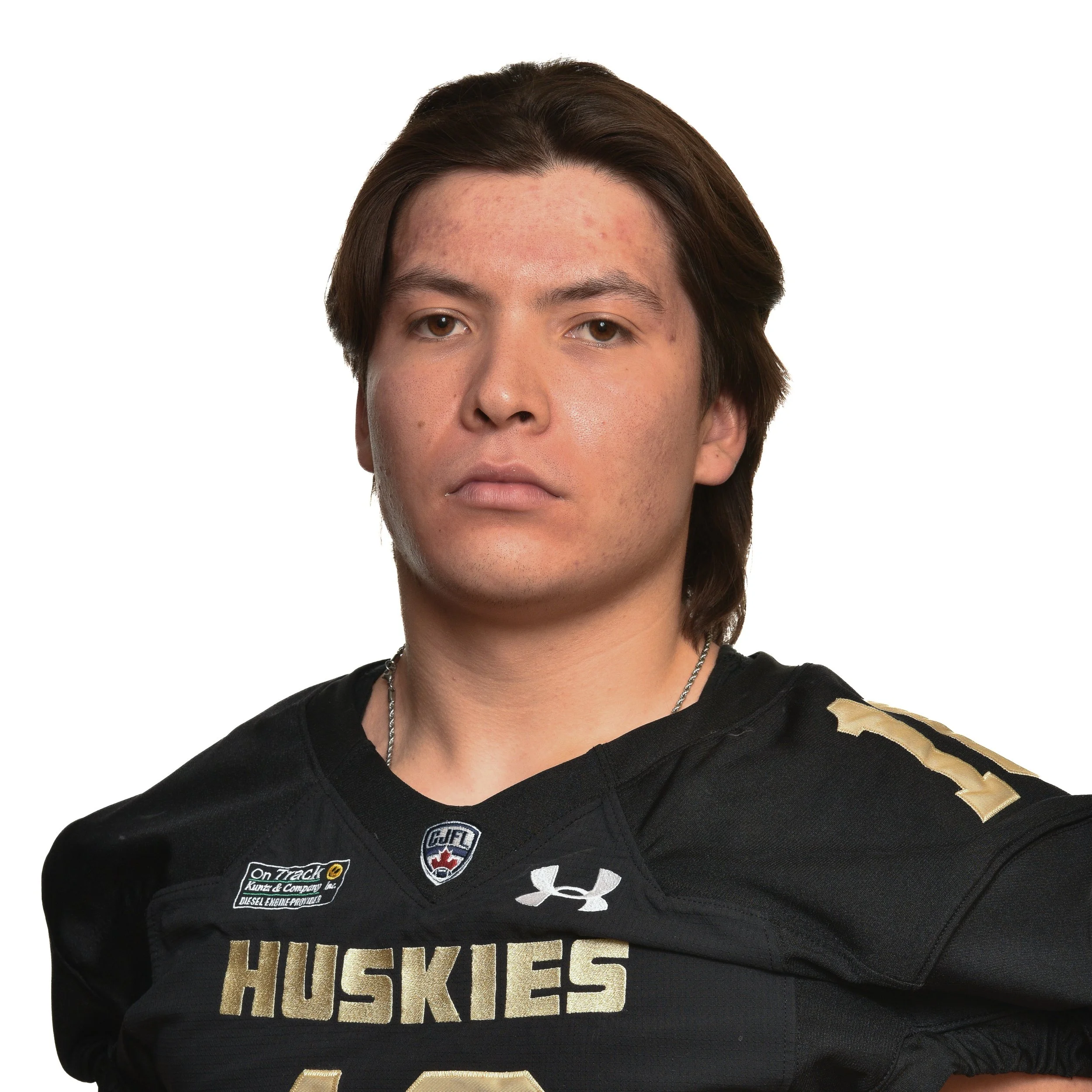 Young man with shoulder-length brown hair wearing a black hockey jersey with "HUSKIES" logo and multiple patches, looking at the camera against a plain background.