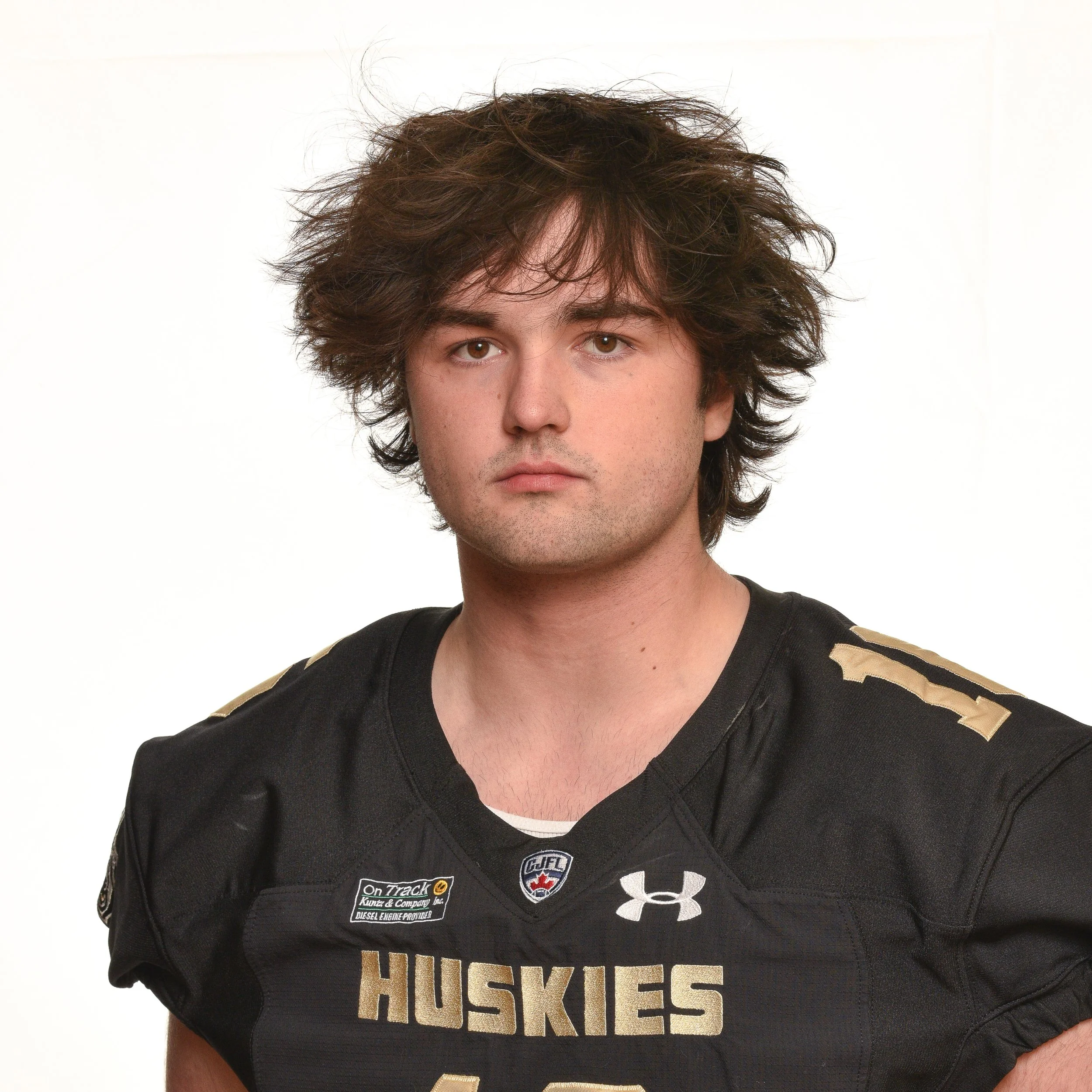 Young man in a black football jersey with Husky and Under Armour logos, looking serious, against a black background.