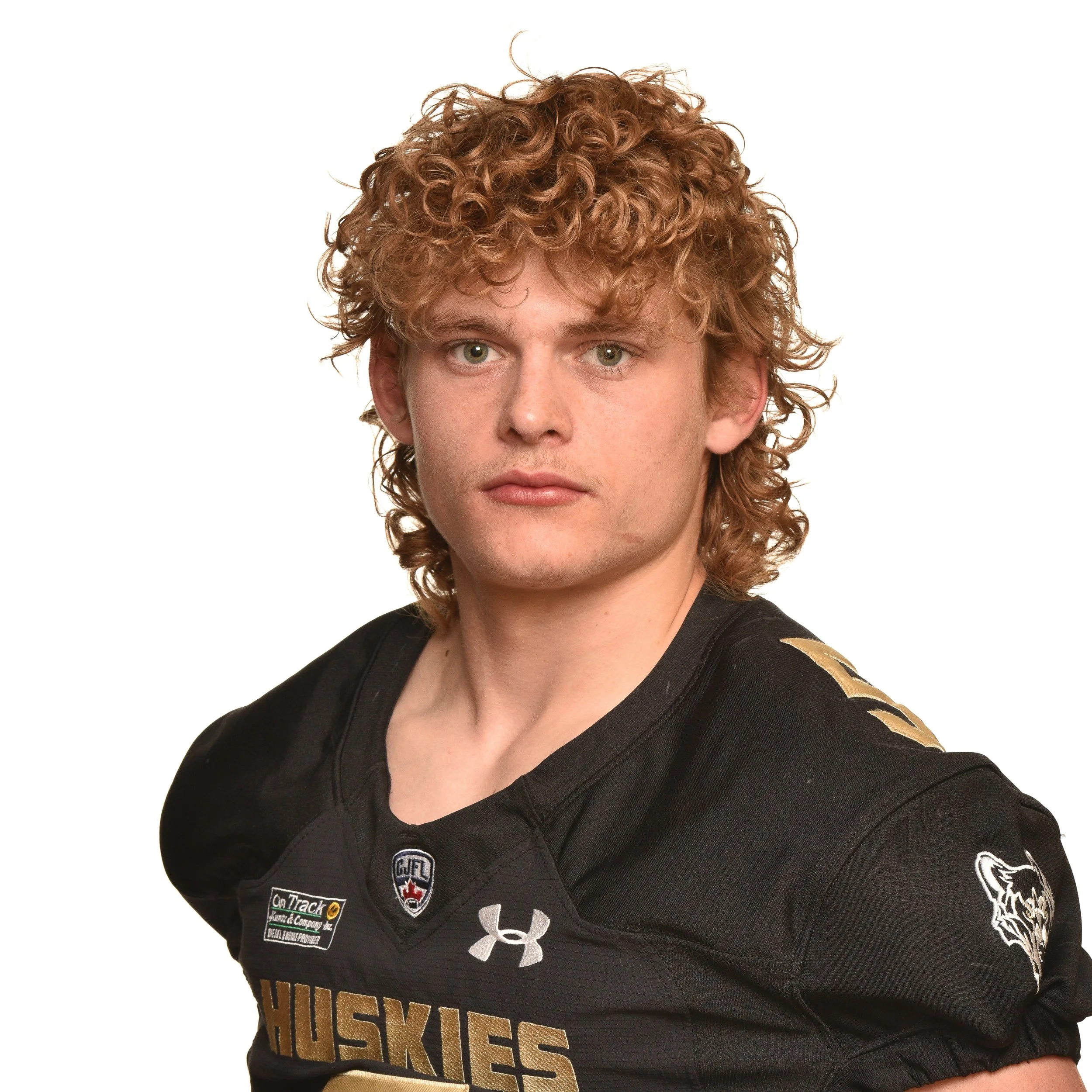 A young male football player with curly red hair wearing a black and gold University of Edmonton Huskies football jersey, standing with hands behind back, looking at the camera.
