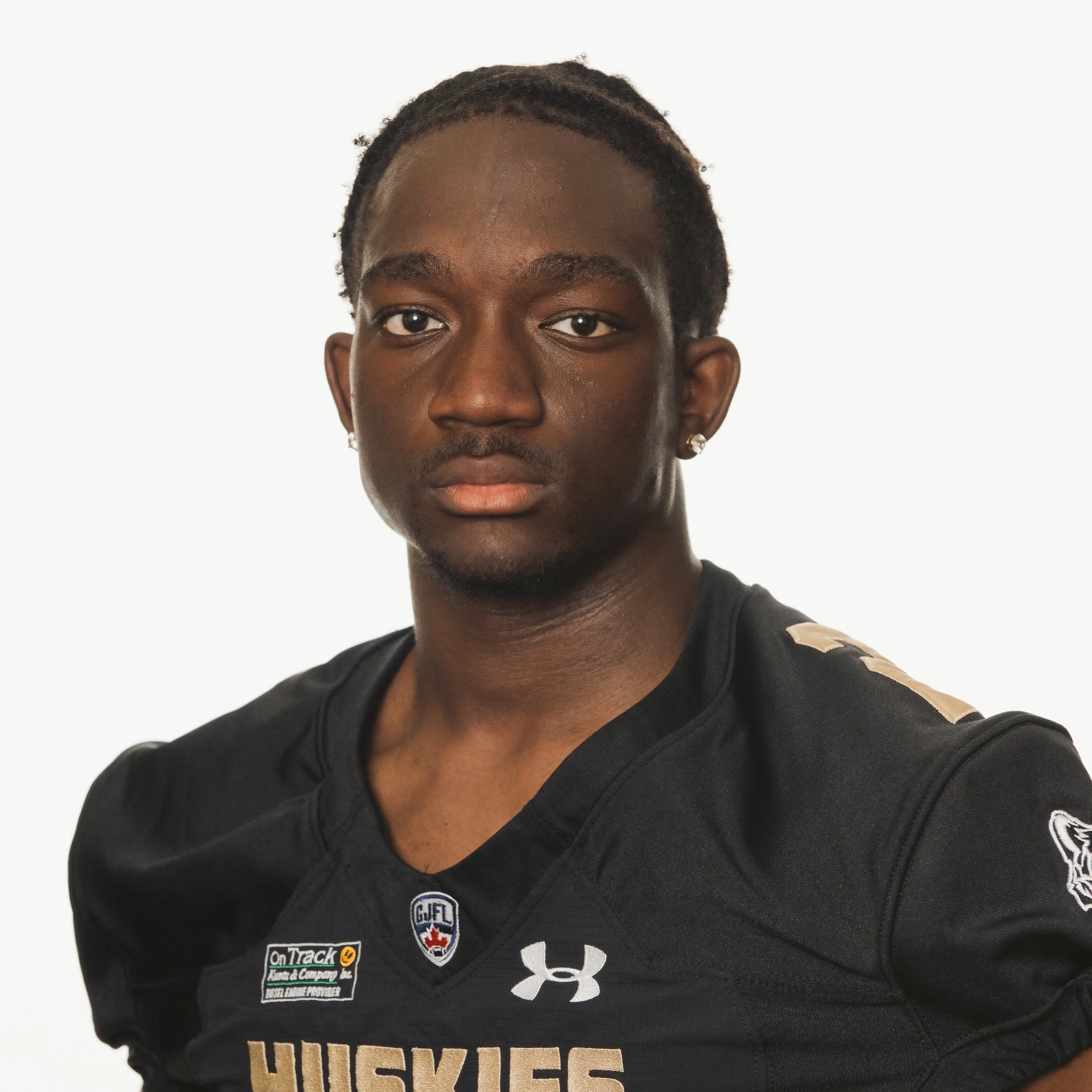 A young African American male football player wearing a black jersey with gold letters and numbers, with the number 24, standing with one hand on his hip. The jersey features the word 'HUSKIES' and various sponsor patches.
