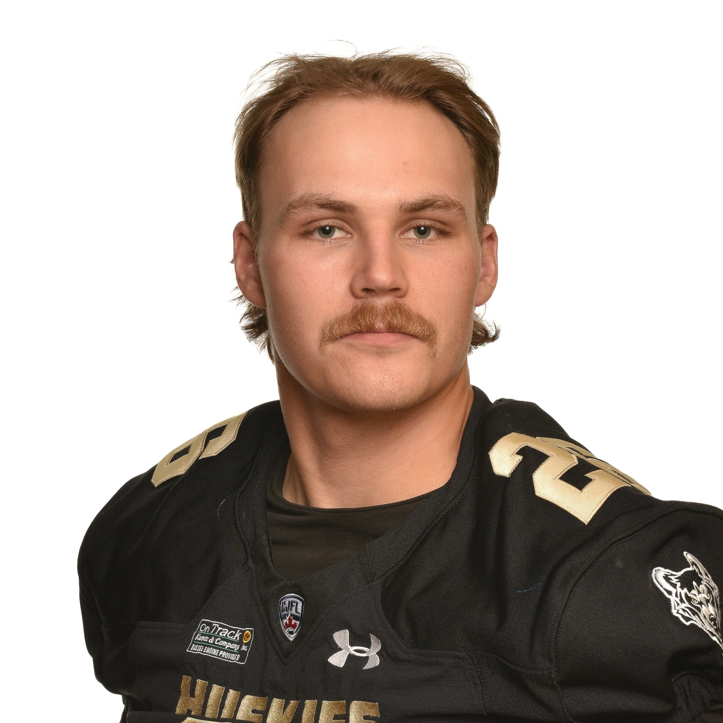 Young man with light skin, long wavy brown hair, and a mustache wearing a black football jersey with team name 'Huskies' and various logos.
