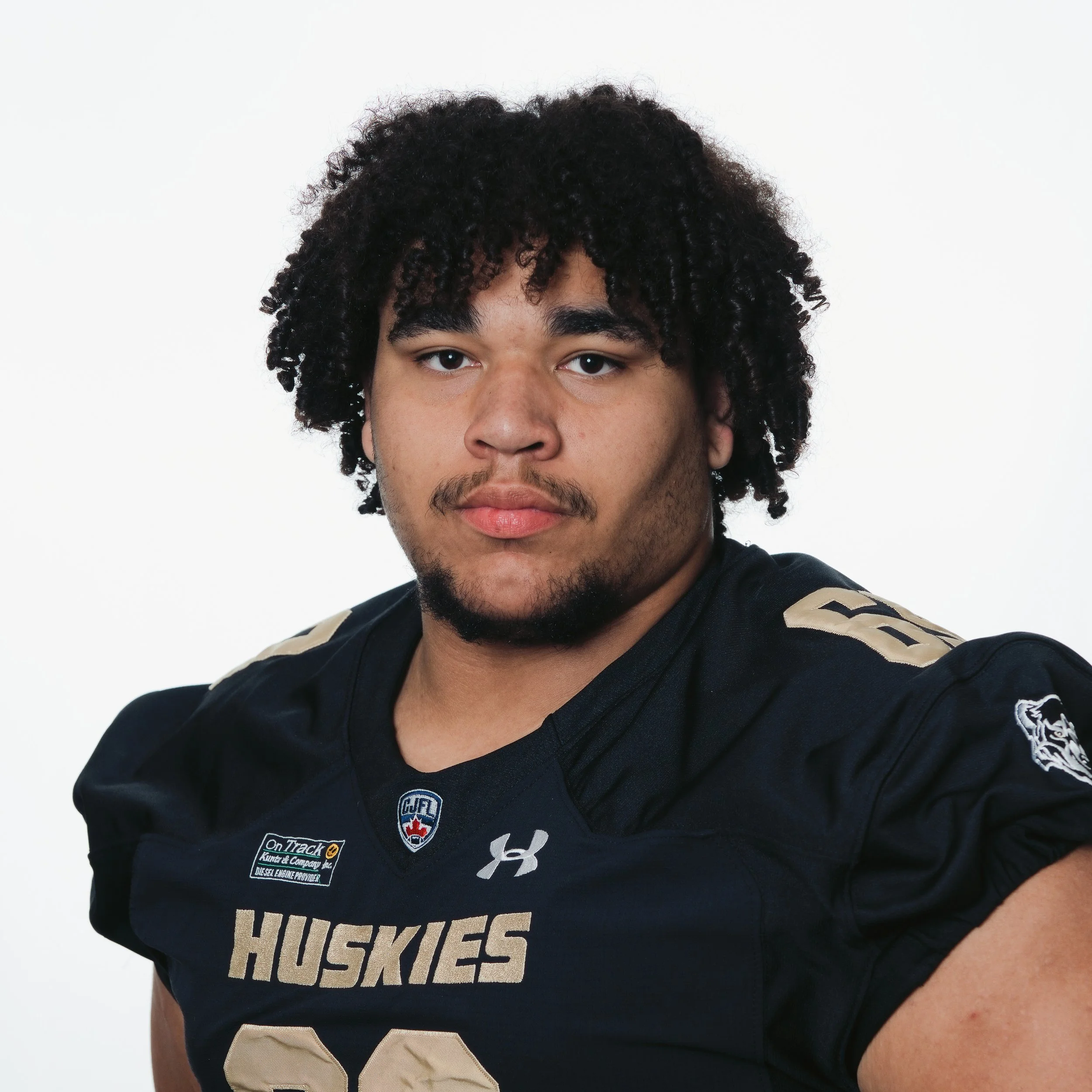 Portrait of a young man with curly brown hair wearing a black football jersey with 'Huskies' written on it.