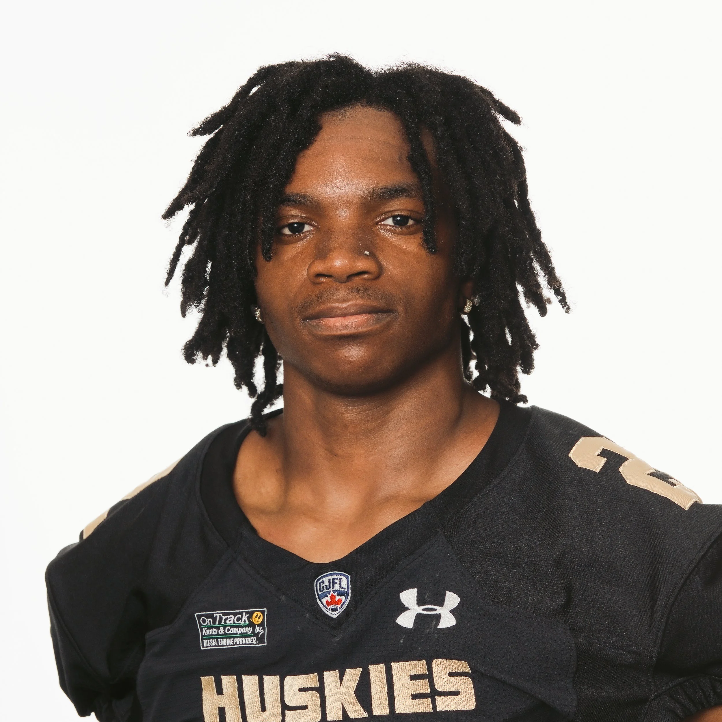 A young African American male football player wearing a black jersey with 'HUSKIES' written on it, a patch with a wolf logo on the shoulder, and a small Canadian Football League logo on the chest. He has short dreadlocks, earrings, and a gold chain, and is posing against a plain background.