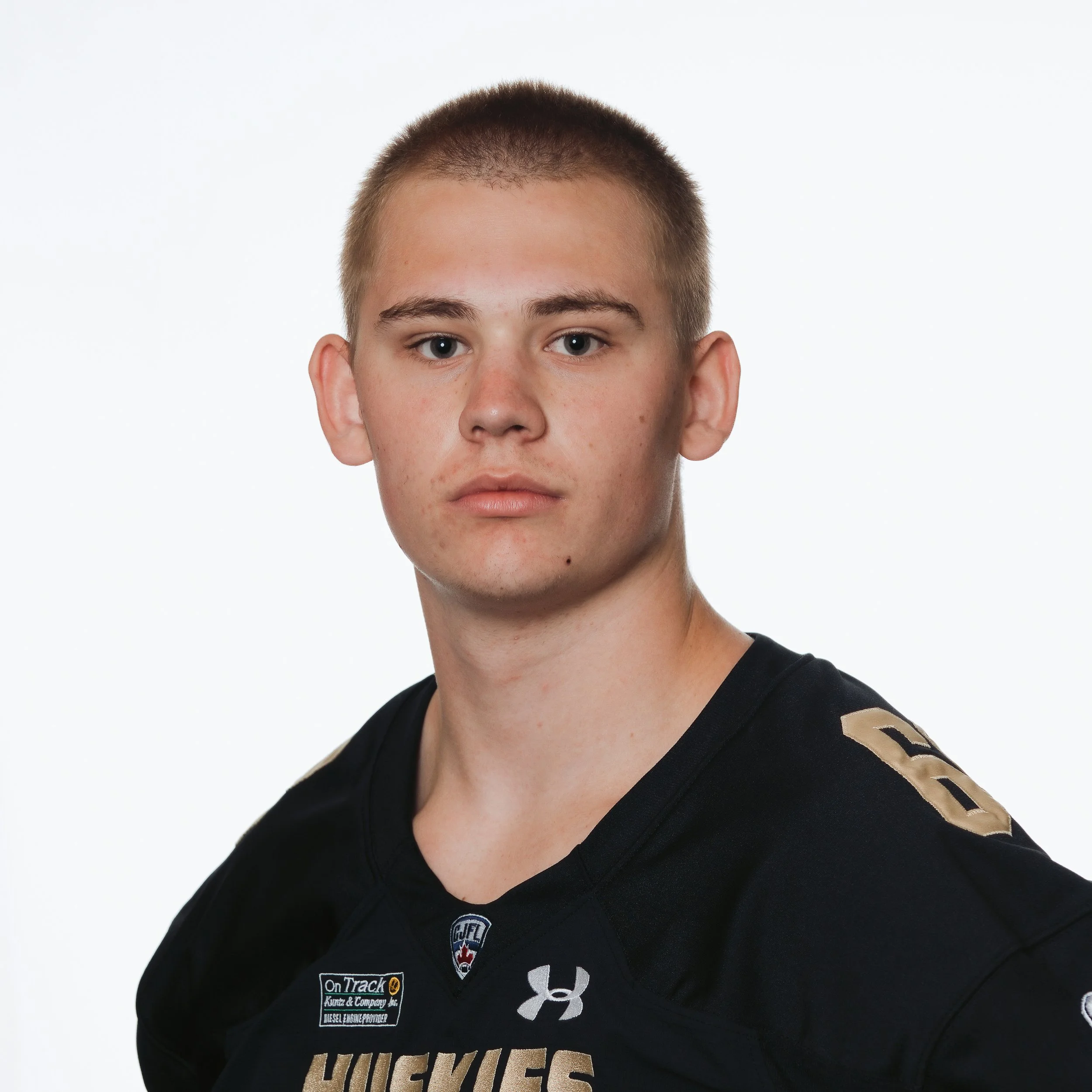 A young male football player with short, reddish hair wearing a black jersey with the word "Huskies" and a wolf logo, standing with a serious expression.