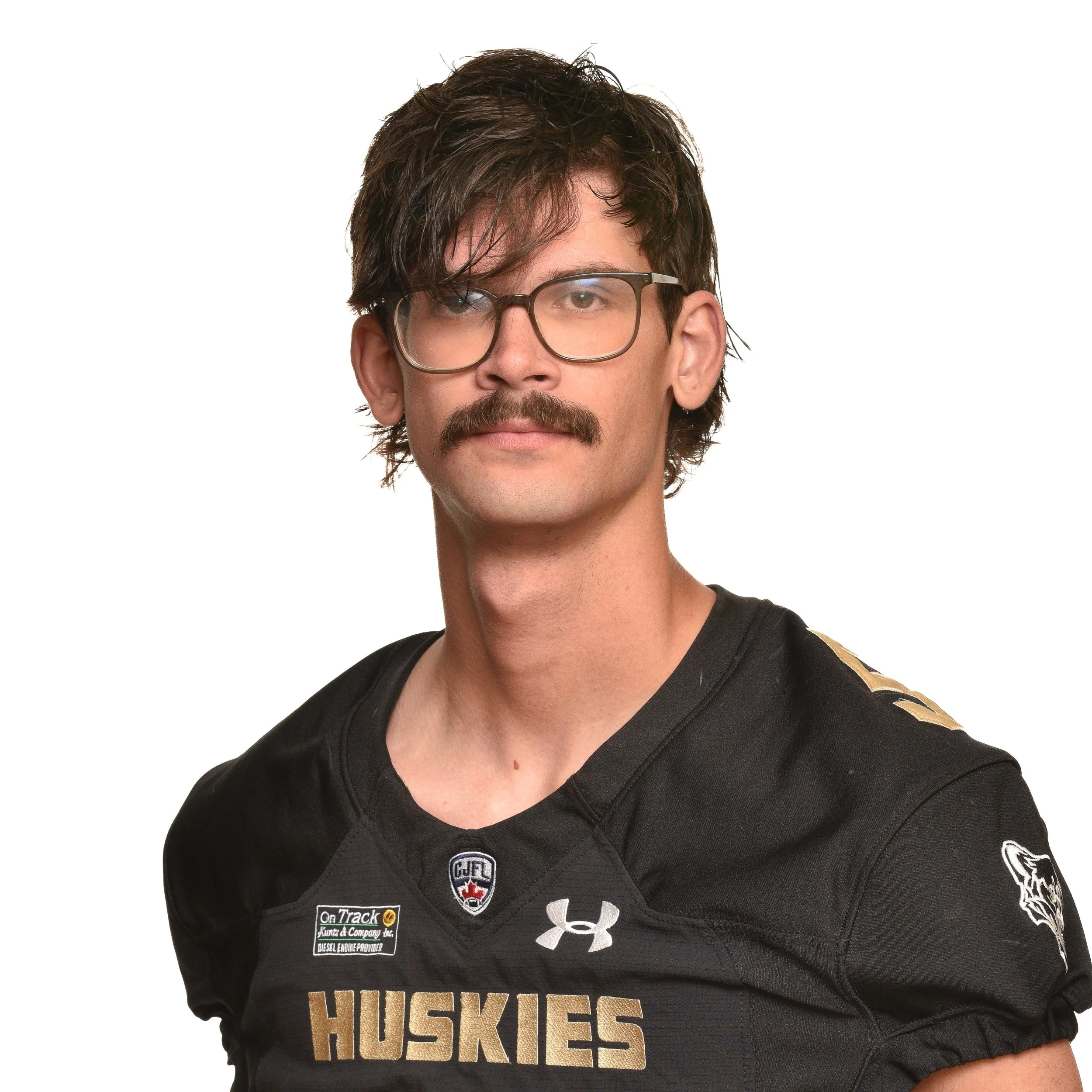 A young man with brown hair, glasses, and a mustache wearing a black football jersey with the word 'HUSKIES' and logos, standing against a black background.