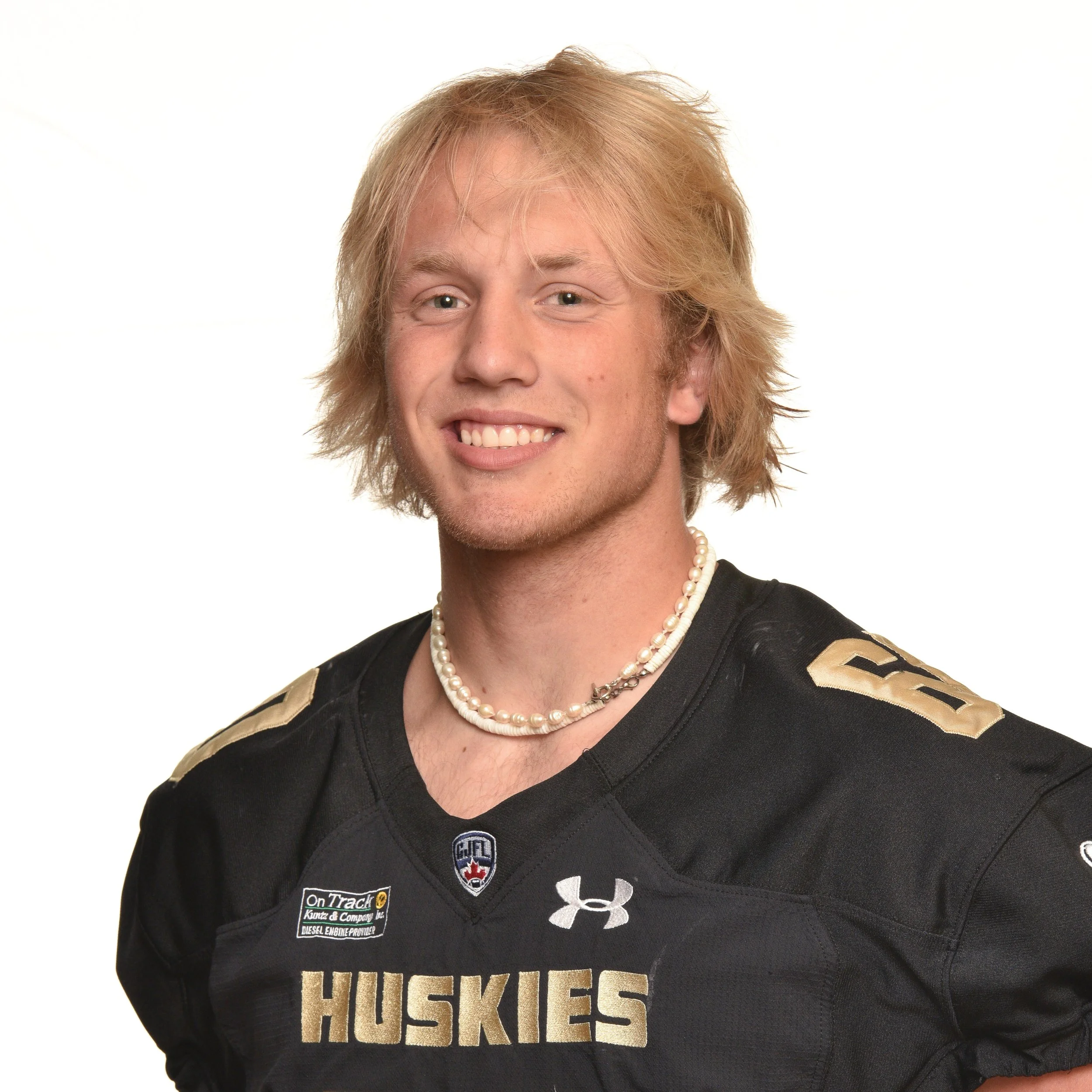 Young man in a black Huskies football jersey with shoulder patches, wearing a beaded necklace, smiling, with shoulder-length blond hair.