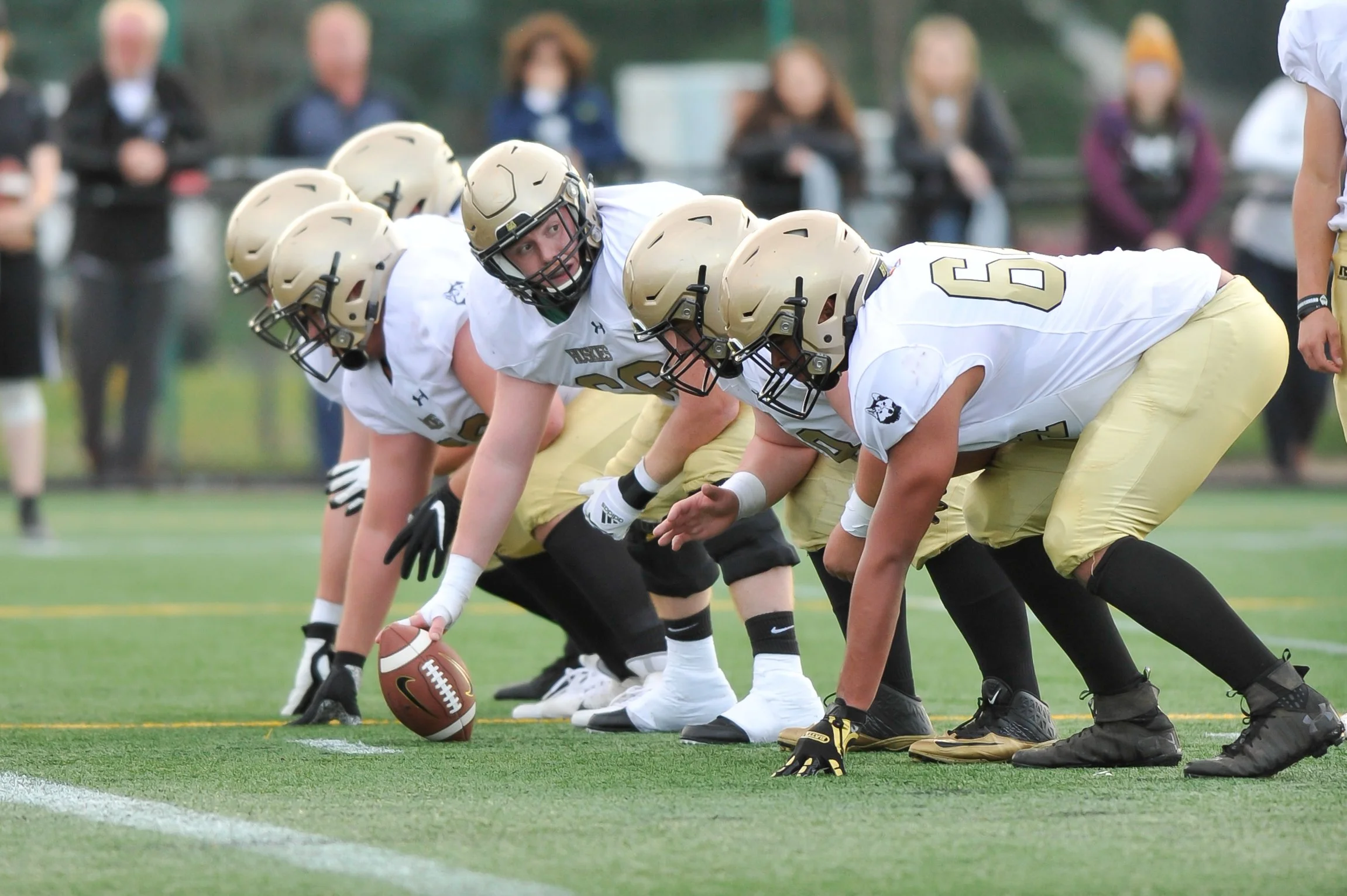 Edmonton Huskies football team line of scrimmage with players in black uniforms and gold helmets crouching in a ready stance on a football field.