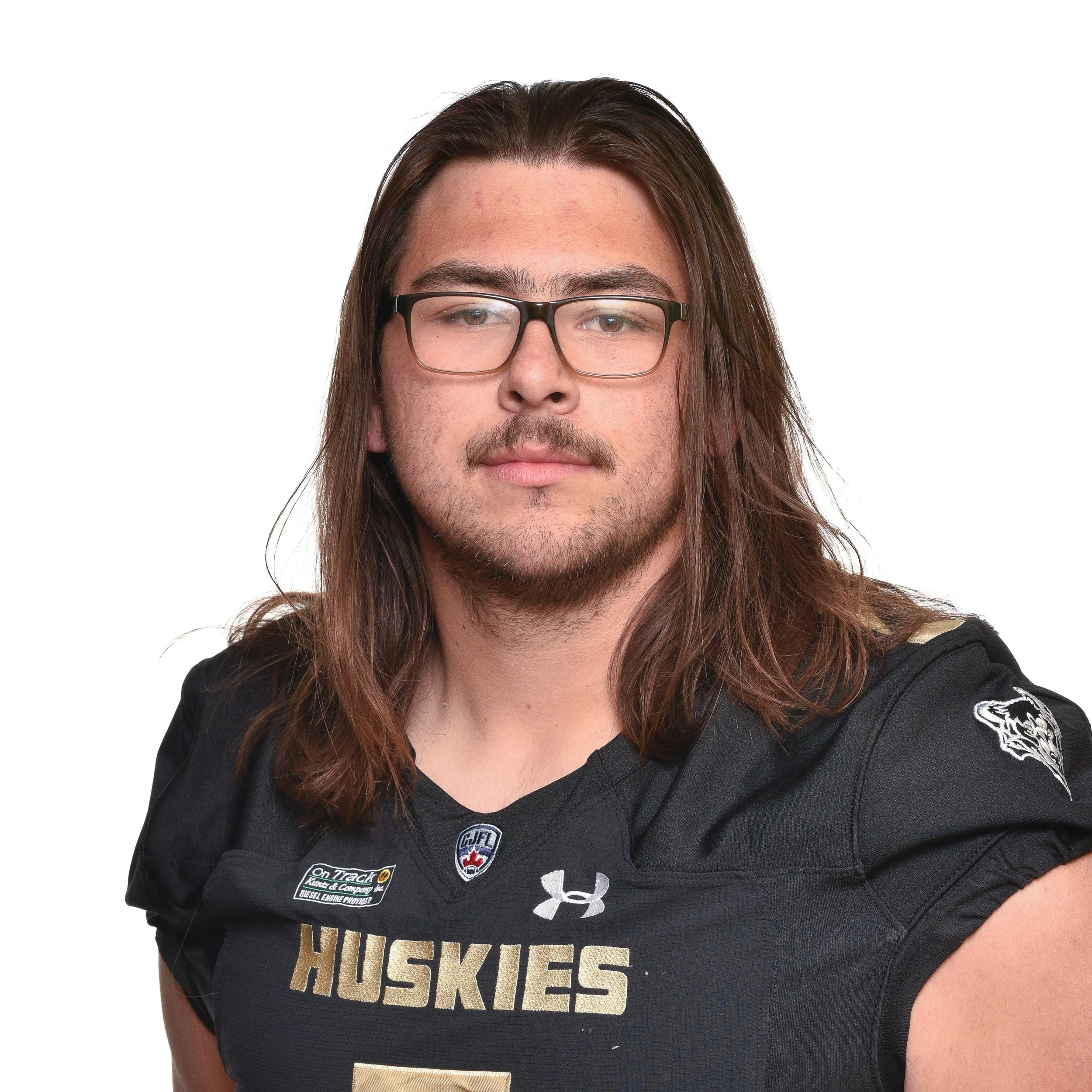 Young man with long brown hair, wearing glasses and a black football jersey with the word 'HUSKIES' on it and team logos, standing against a plain background.