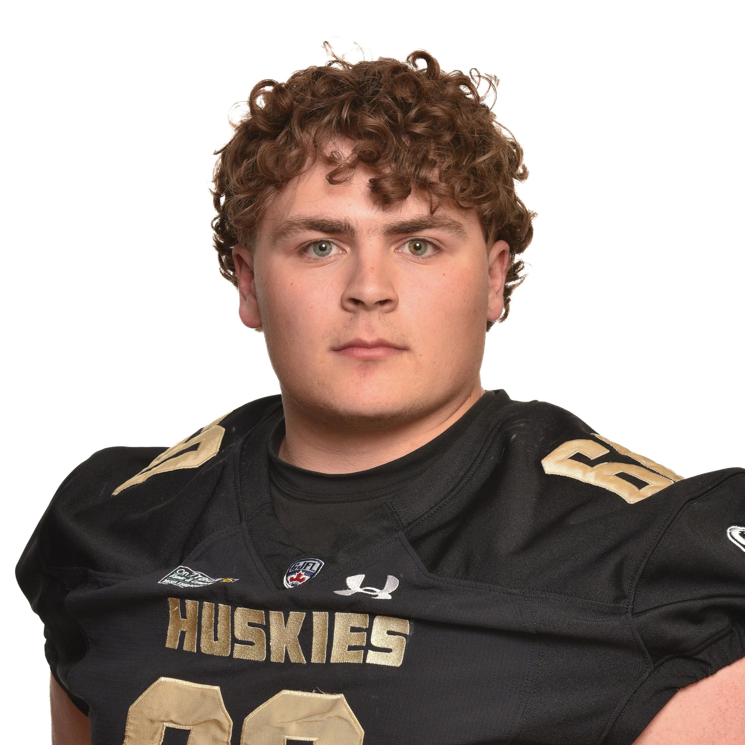 A young man with curly brown hair wearing a black football jersey with 'HUSKIES' written on it, a shield logo, and various patches.