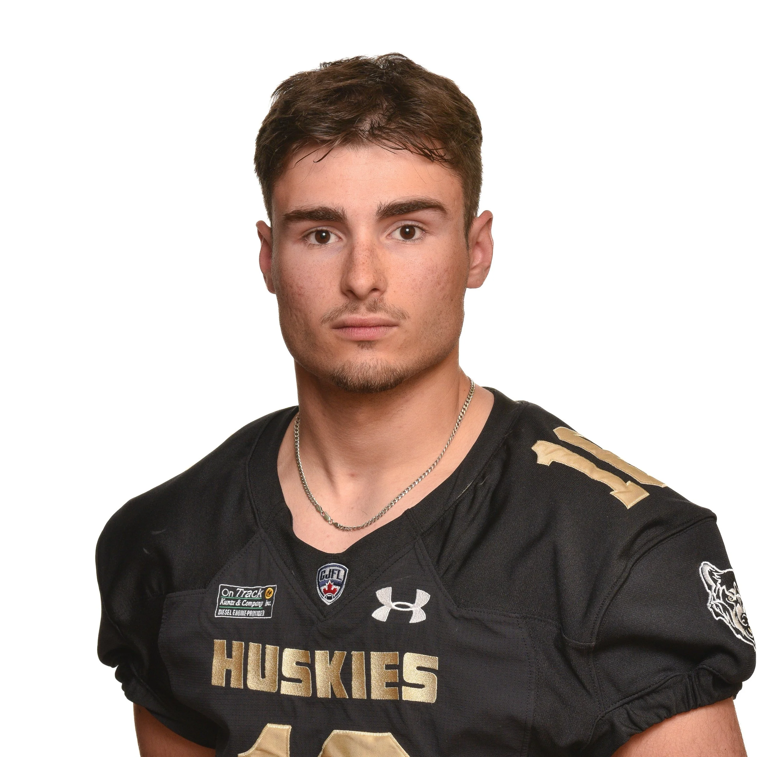A young man with short brown hair, wearing glasses, a black football jersey with the word 'Huskies' on it, and a thin silver chain necklace.