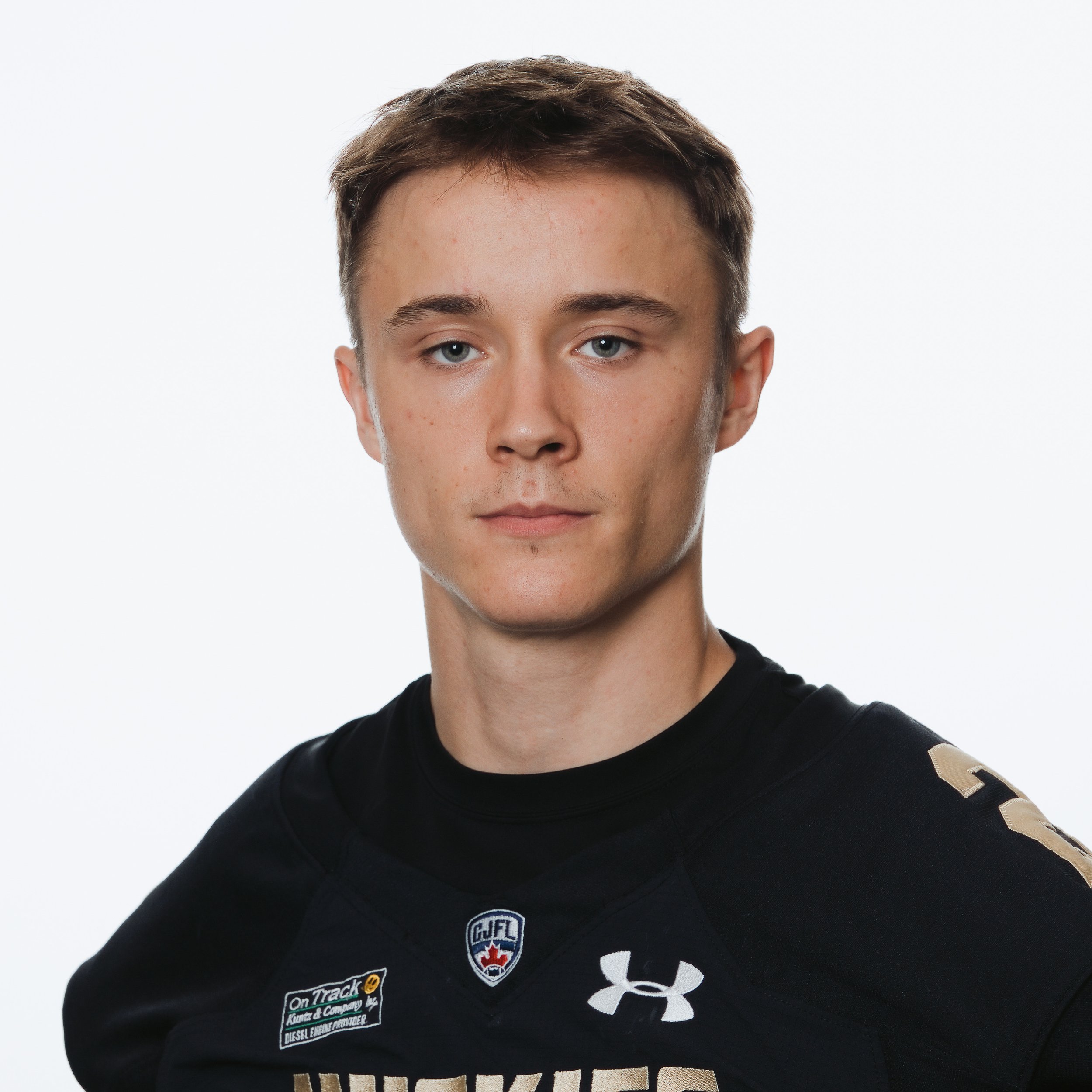 Young man with short brown hair wearing a black football jersey with the word 'HUSKIES' and several logos, including Under Armour and a football league patch.