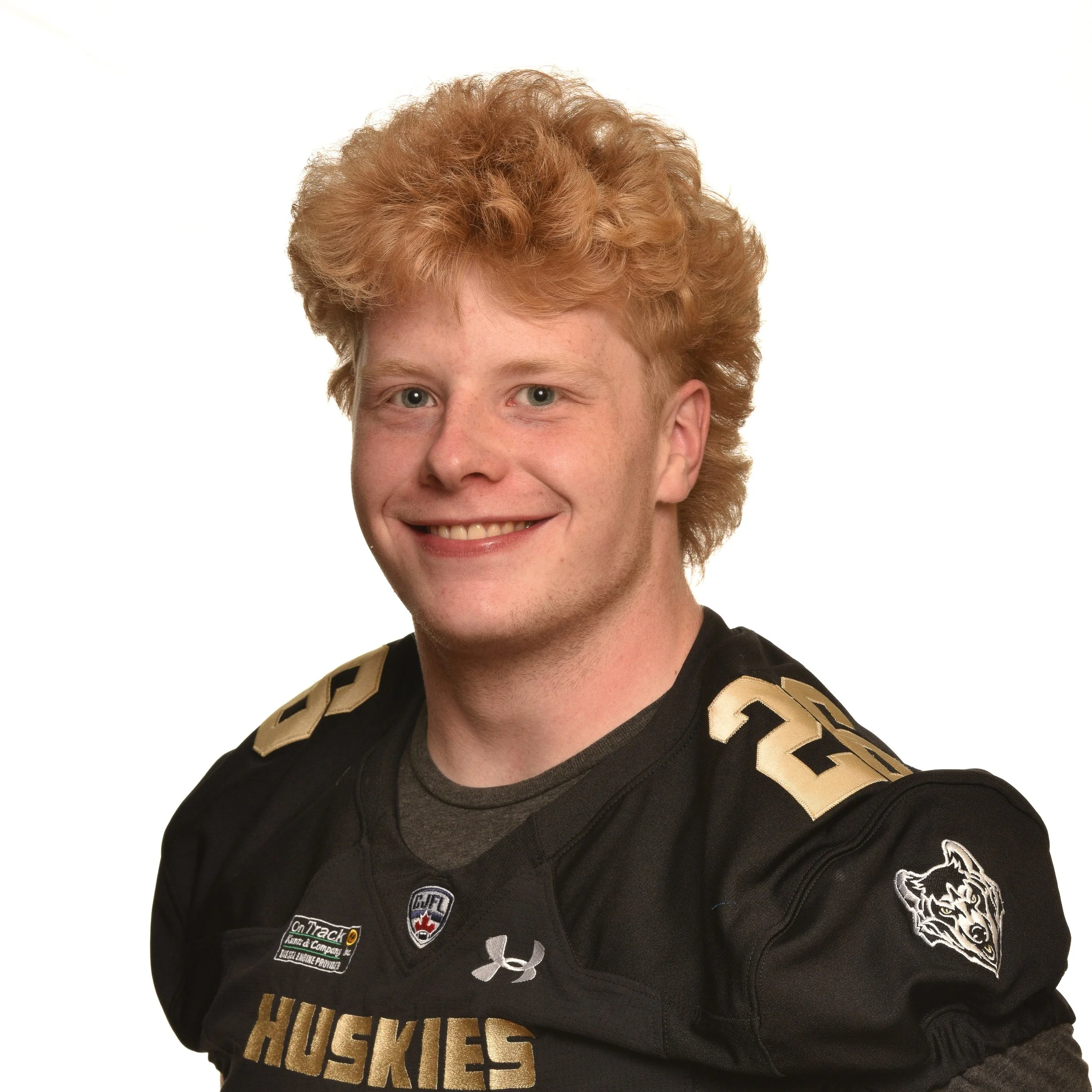 Young man with curly red hair wearing a black football jersey with various patches, including a Husky logo, standing against a black background.