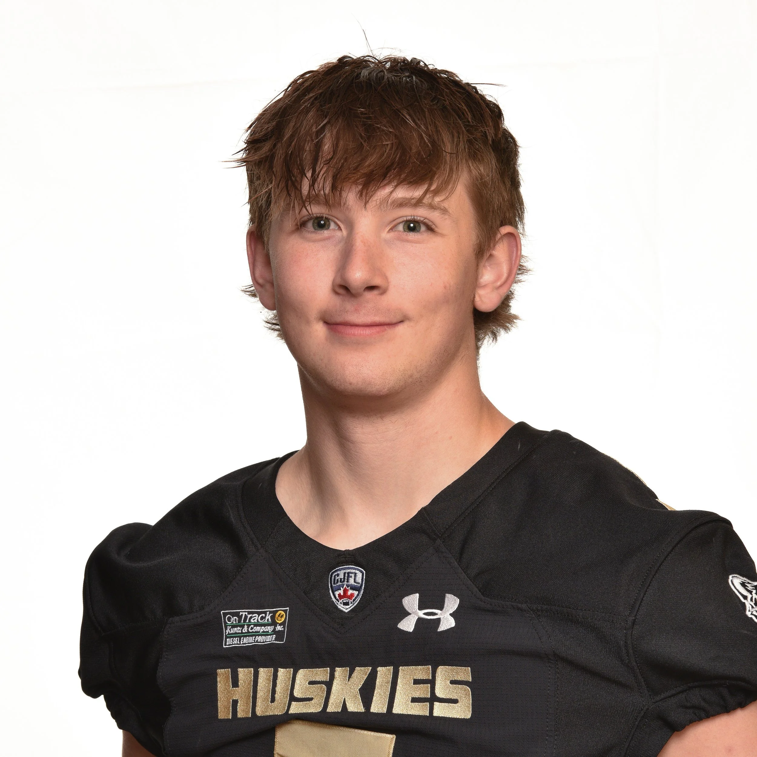 A young man with red hair wearing a Black Under Armour hockey jersey with 'HUSKIES' written across the chest, posing for a portrait.