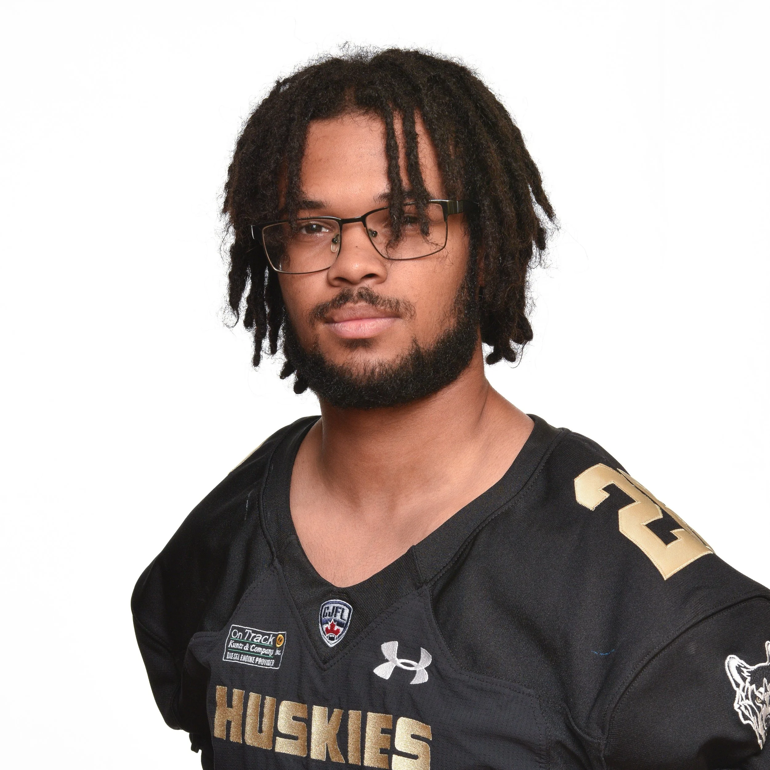 Portrait of a young man with an afro hairstyle wearing a black football jersey with 'Huskies' written on it, and patches including a wolf logo and sponsor logos, against a white background.
